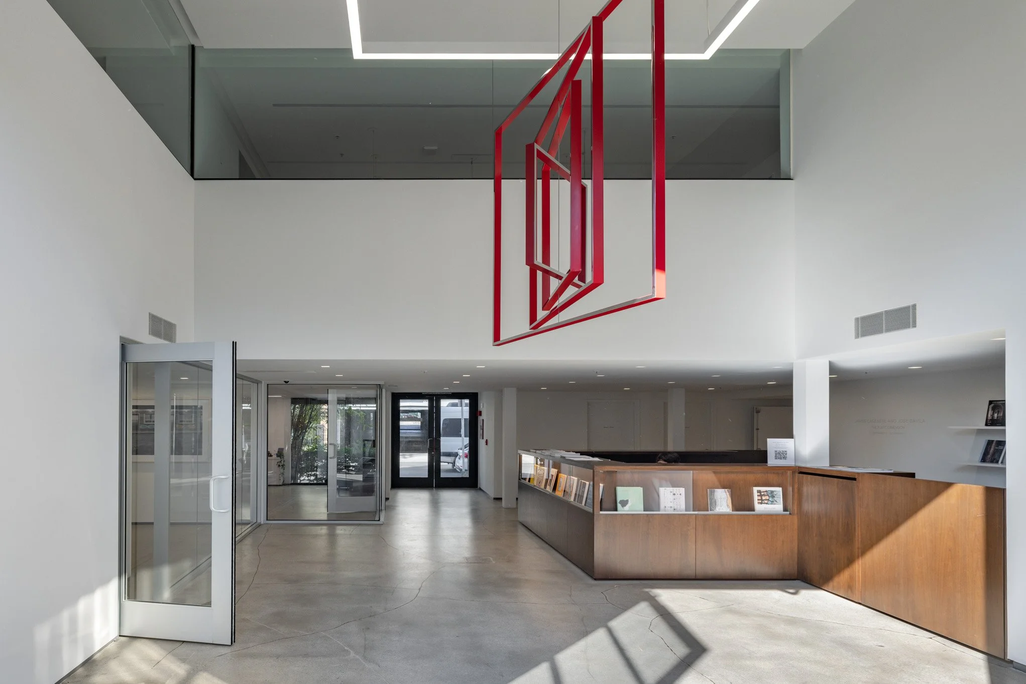Interior of a modern building lobby with white walls, a wooden reception desk, glass doors, and a decorative red metal art piece hanging from the ceiling.