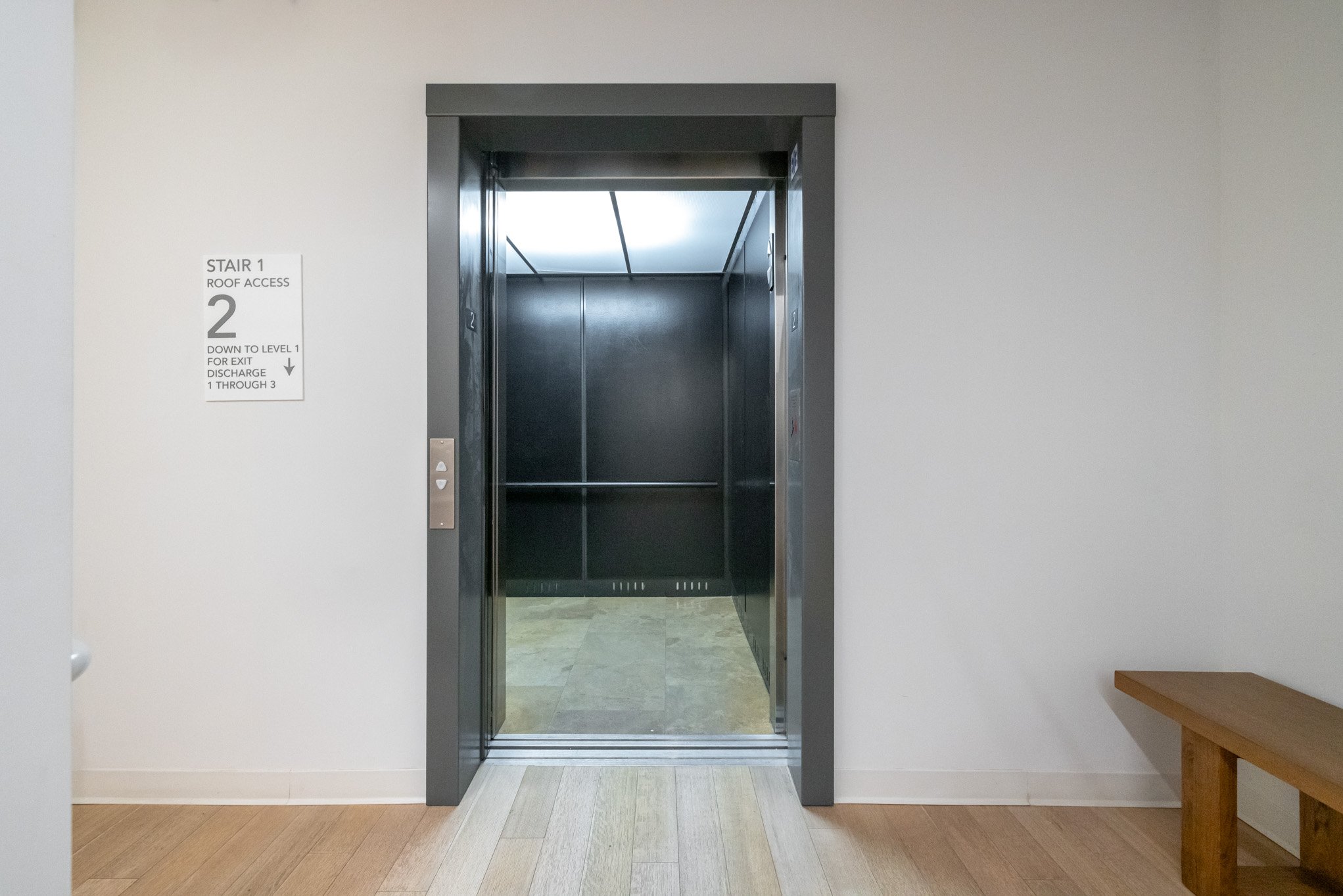 Elevator with open doors in a modern building, white wall on the left with a sign indicating stairs and floor information, wooden bench on the right.