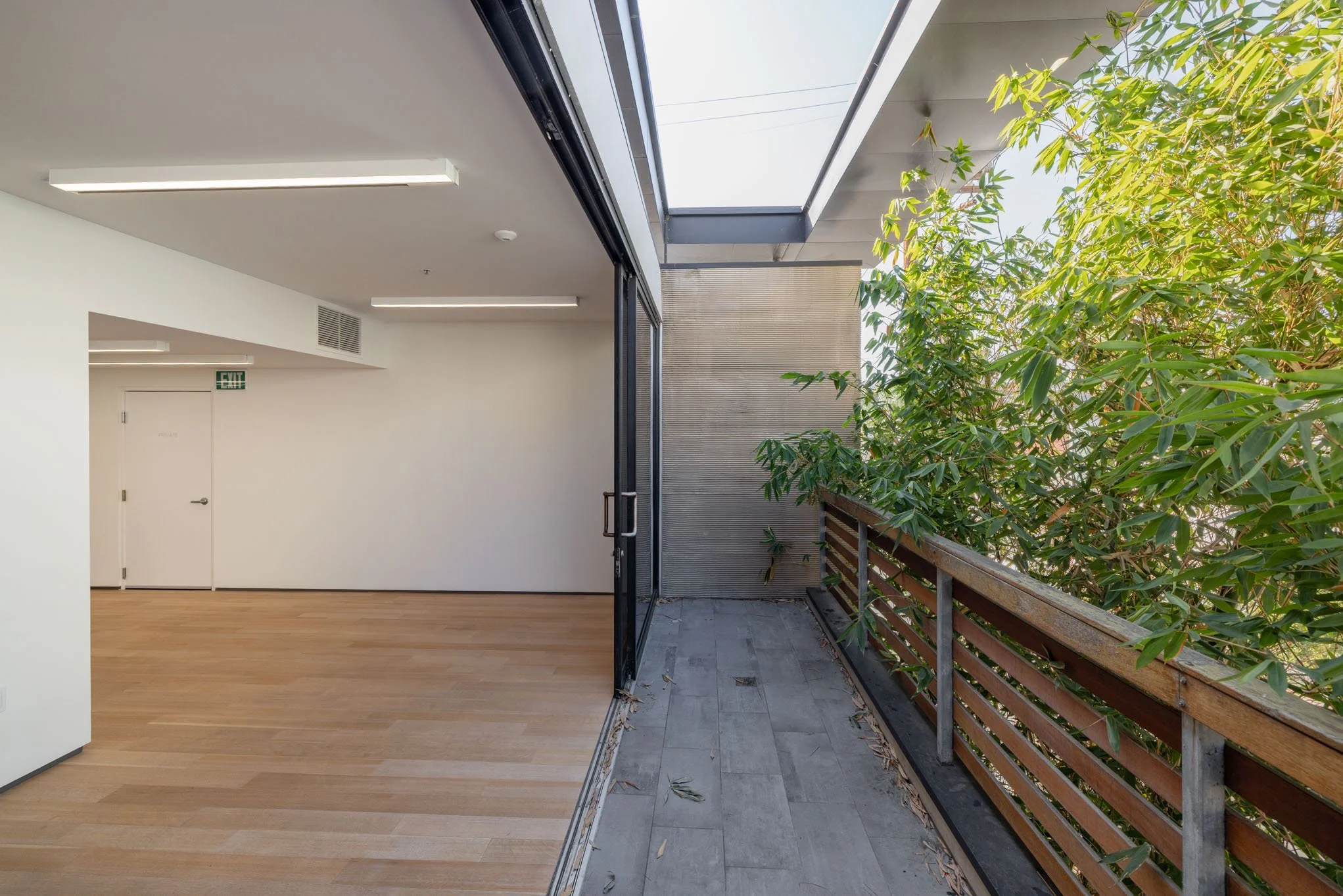 Interior view of a modern apartment or office space with hardwood floors, white walls, and a balcony with a wooden railing and green foliage outside.