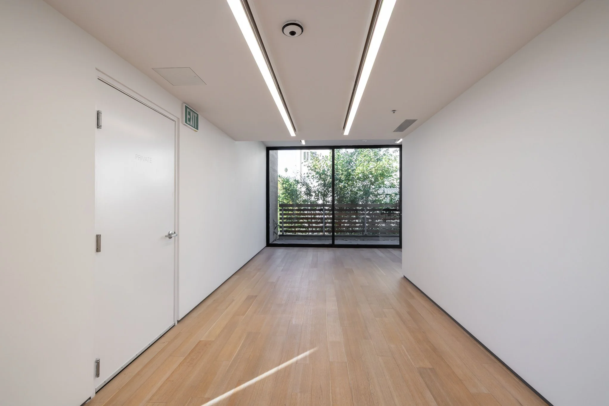 Empty hallway with white walls, wood flooring, large glass sliding doors at the end, and ceiling lights.
