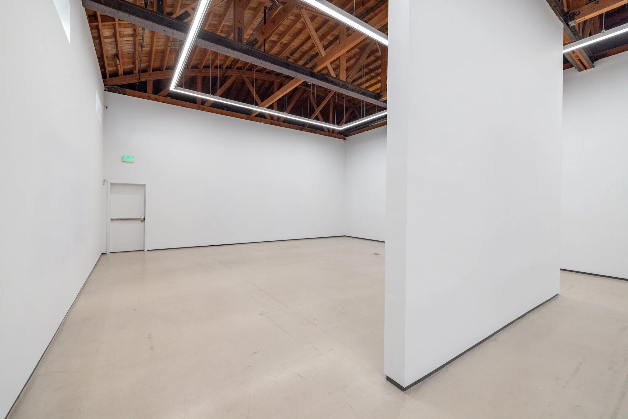 Empty white-walled room with a high exposed wooden ceiling, modern linear lighting, and a door with a green exit sign.