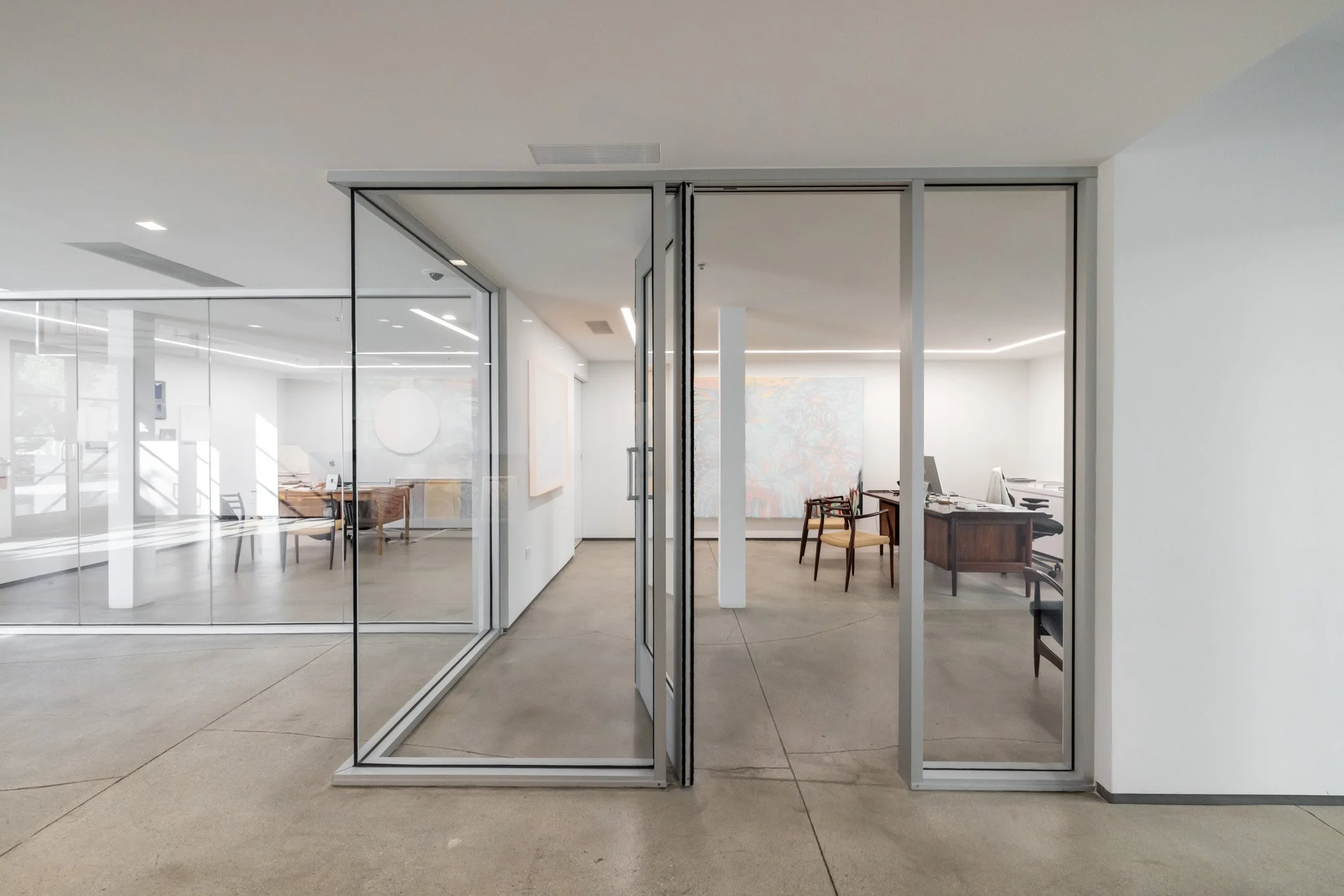 Office space with glass walls and doors, featuring minimalist decor, desks, chairs, and abstract art on the walls.