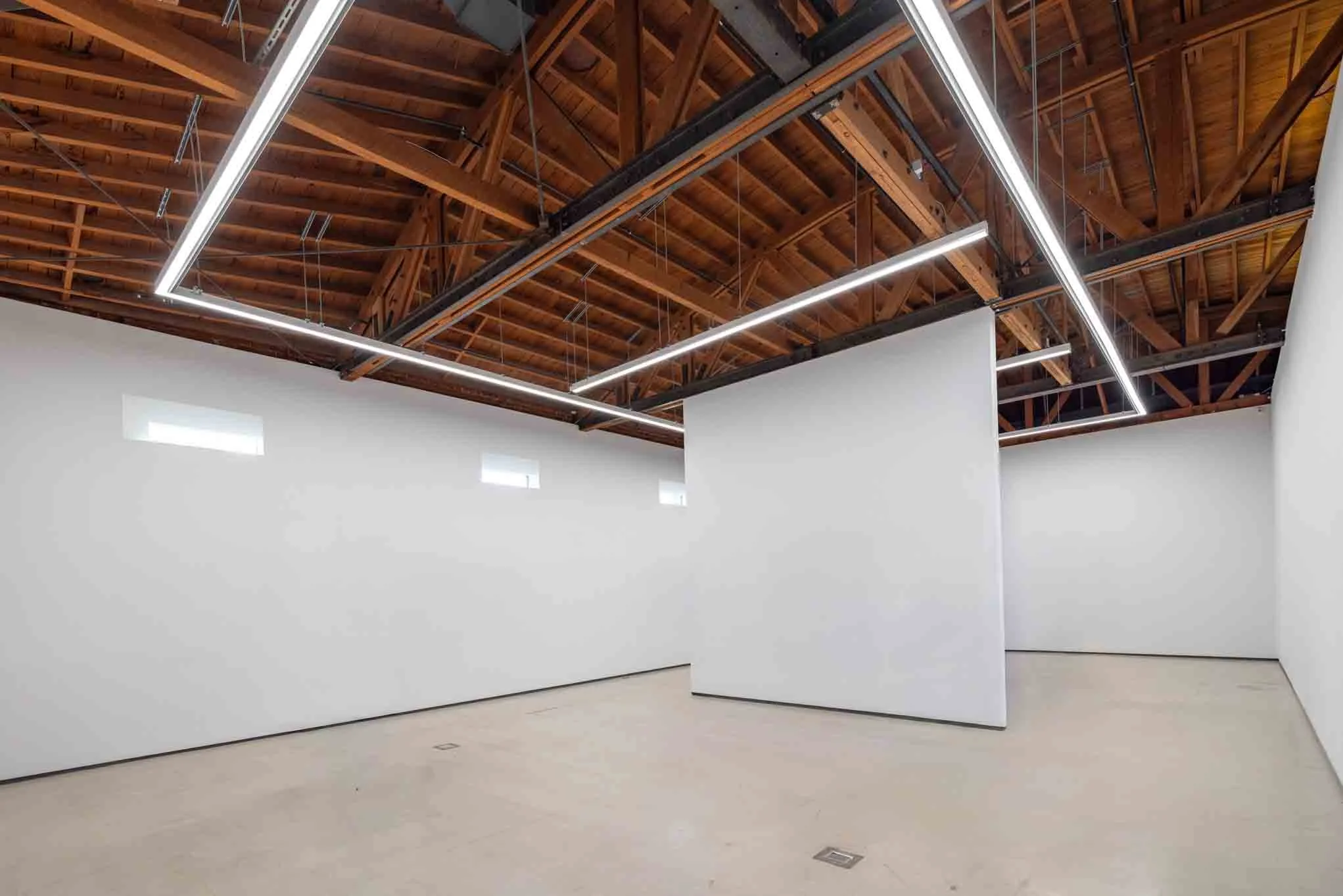 Empty gallery with white walls, exposed wooden ceiling, and modern linear lighting fixtures.