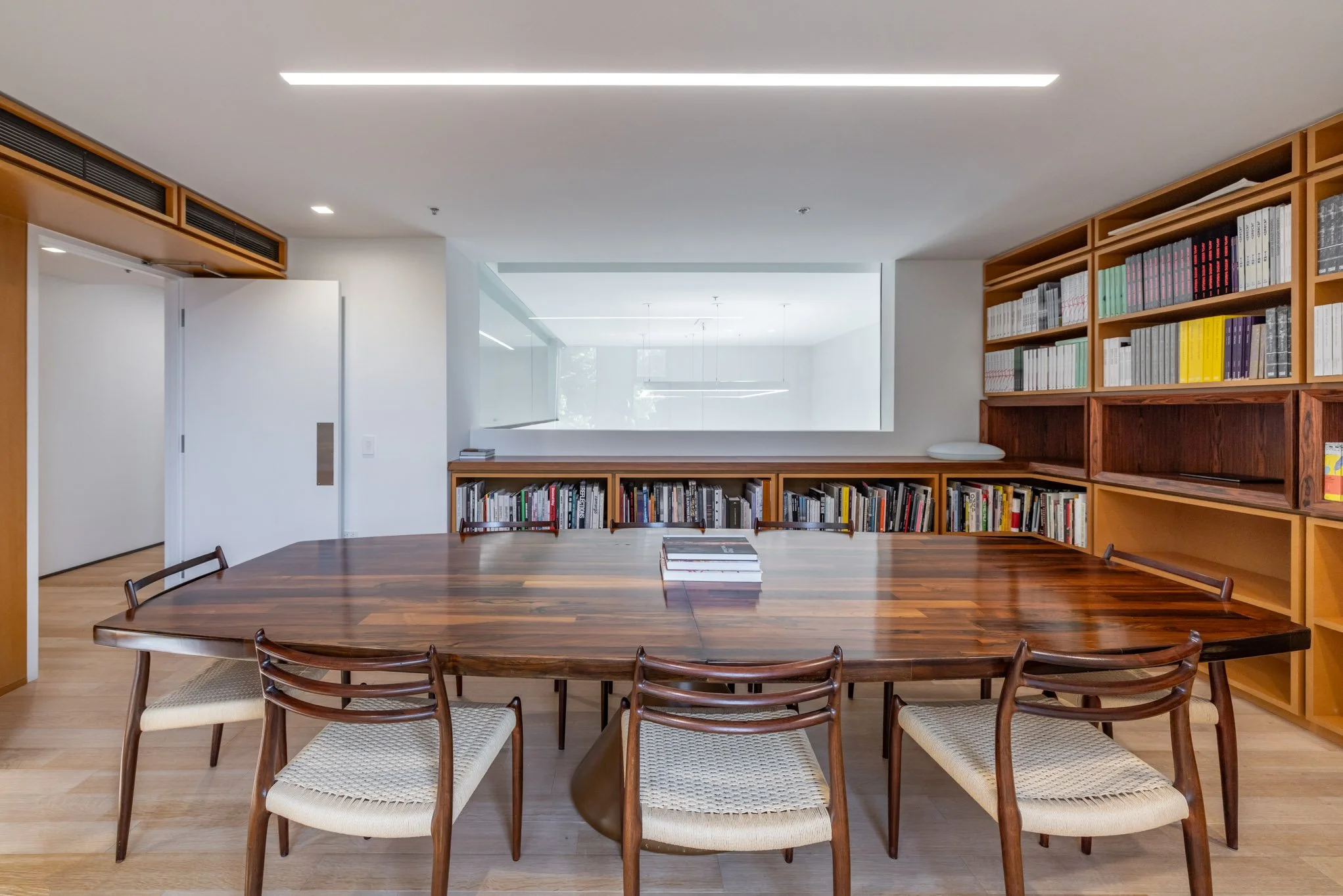 Modern conference room with wooden table and six chairs, bookshelf with books, a large window, and white walls.