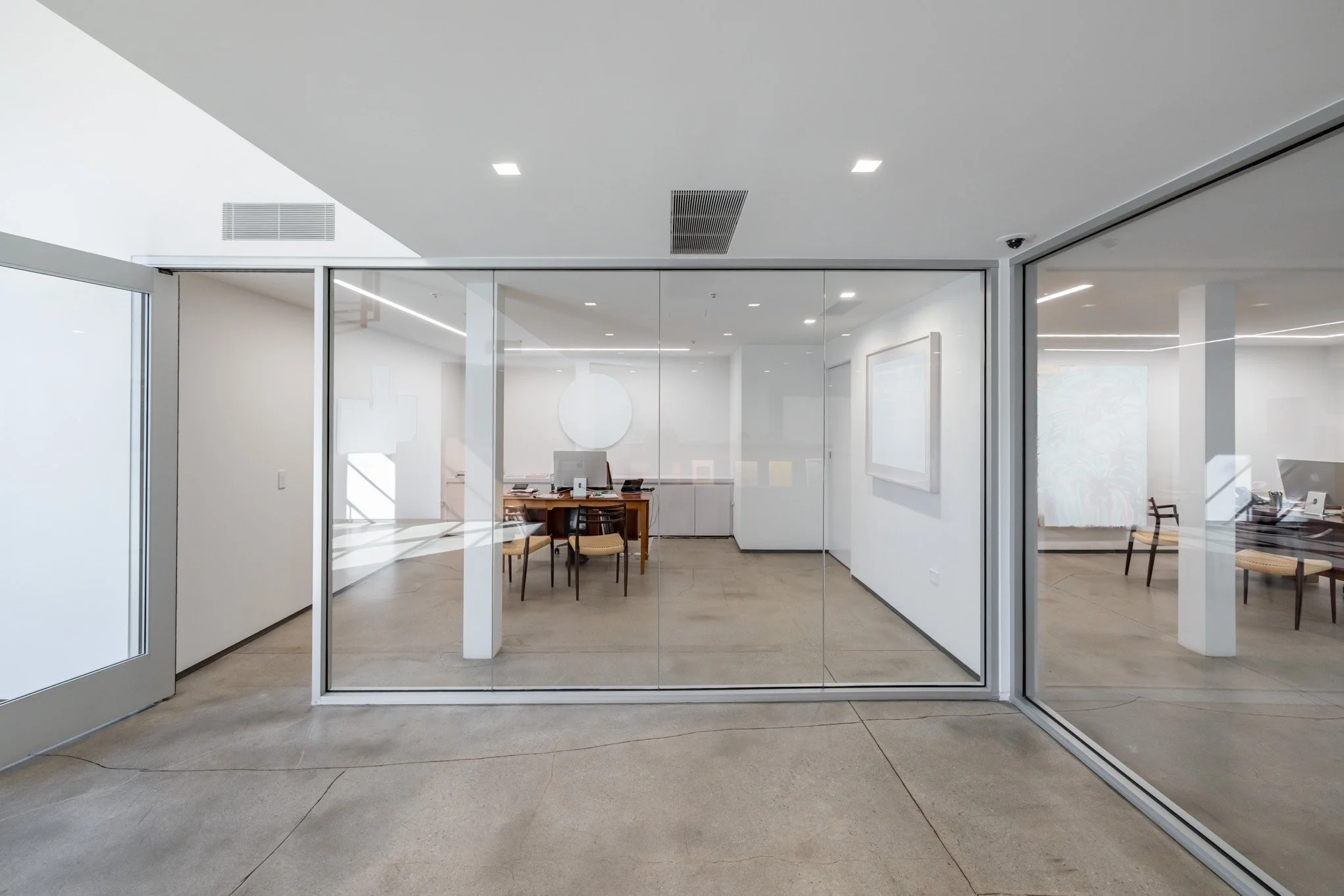 Interior view of an office with glass walls and doors, a desk with an iMac computer, chairs, and minimal decor.