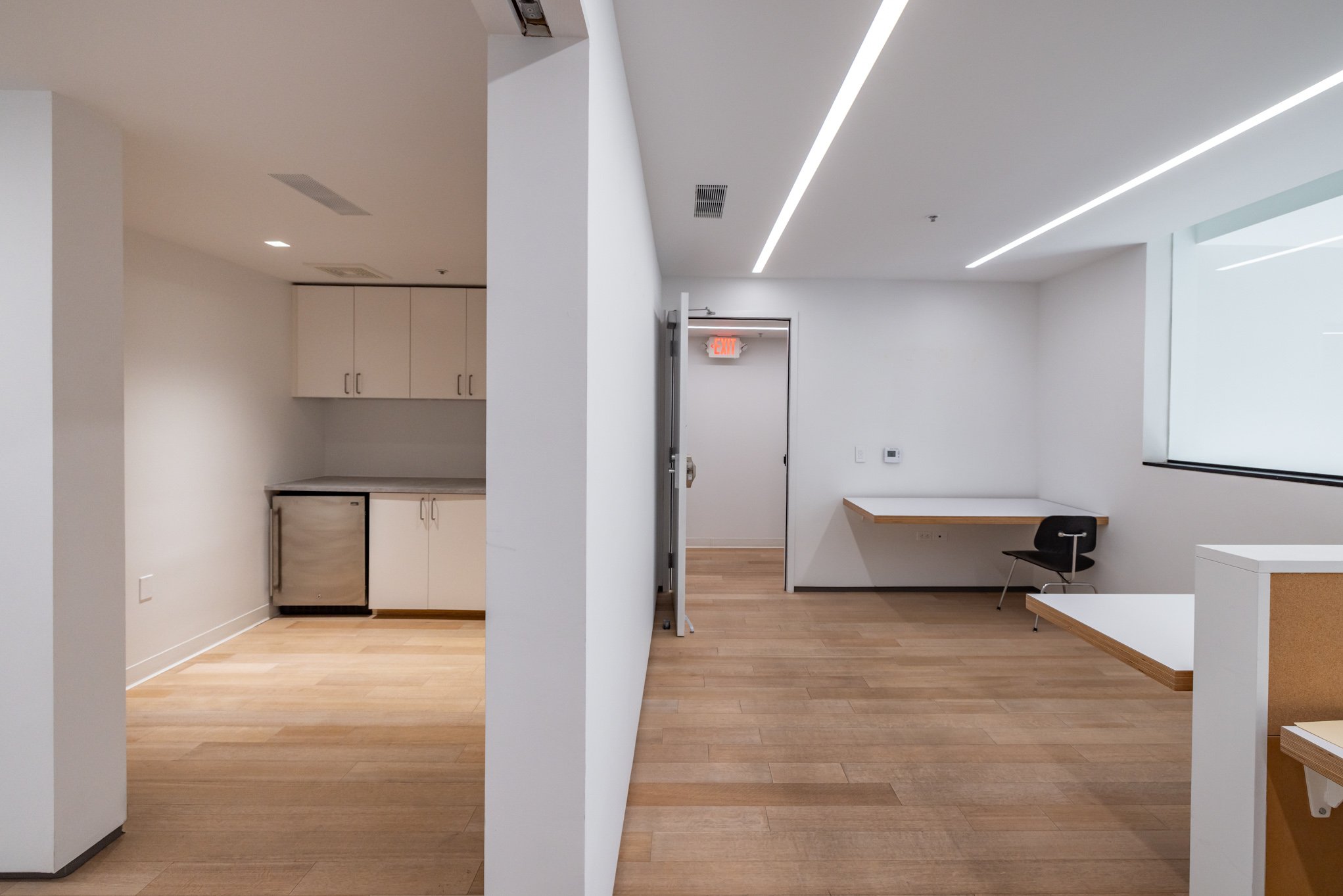 Empty office space with white walls, wood flooring, a small kitchenette to the left, a wall-mounted desk with a chair to the right, and a hallway with an exit sign in the background.