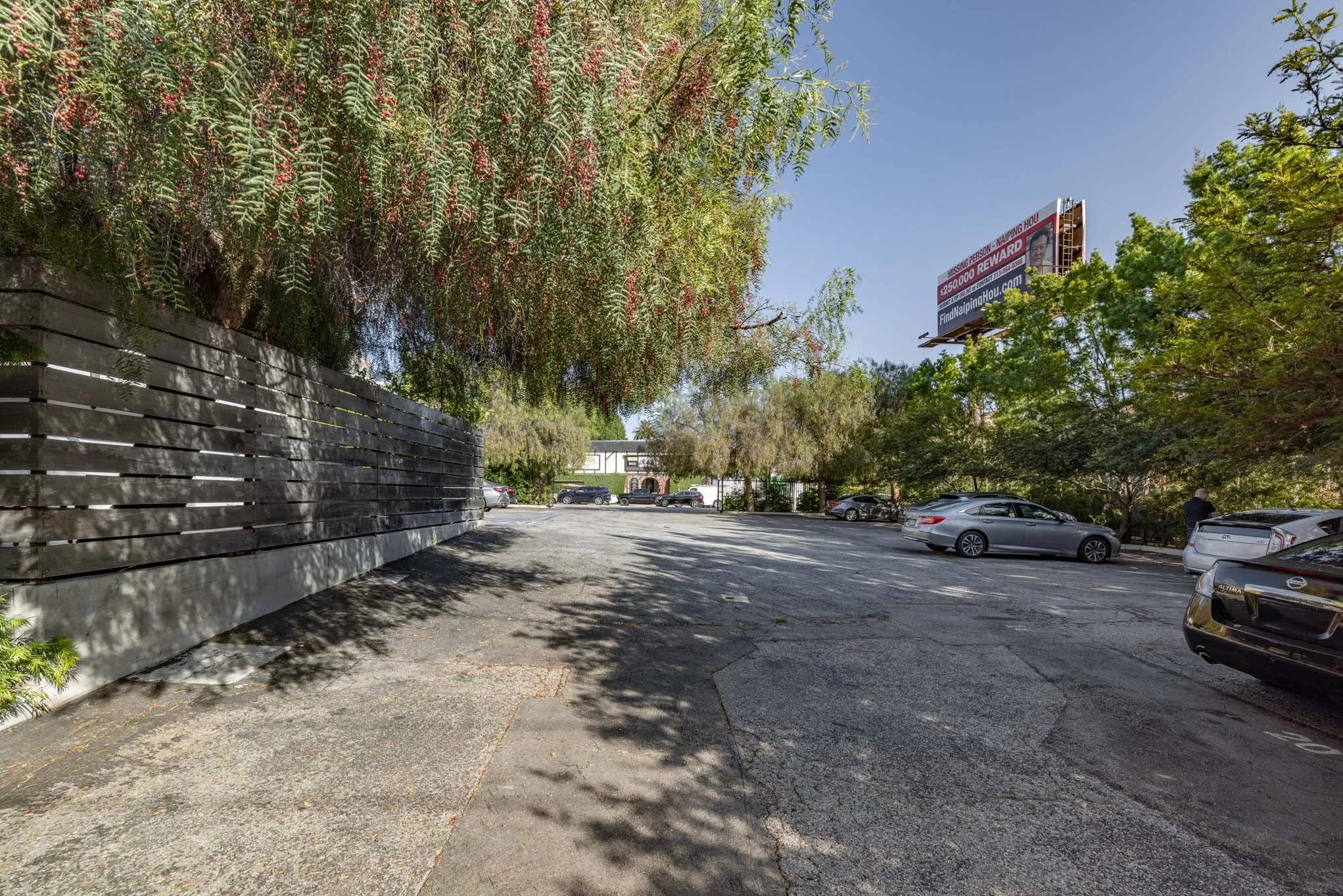 A parking lot with multiple parked cars, surrounded by trees and a wooden fence on the left. A billboard is visible in the background above the trees. The scene is bright with clear skies.