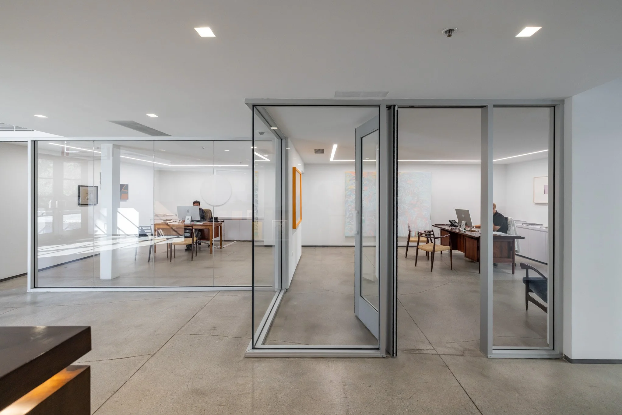 Modern office with glass walls and doors, featuring two employees working at desks inside.