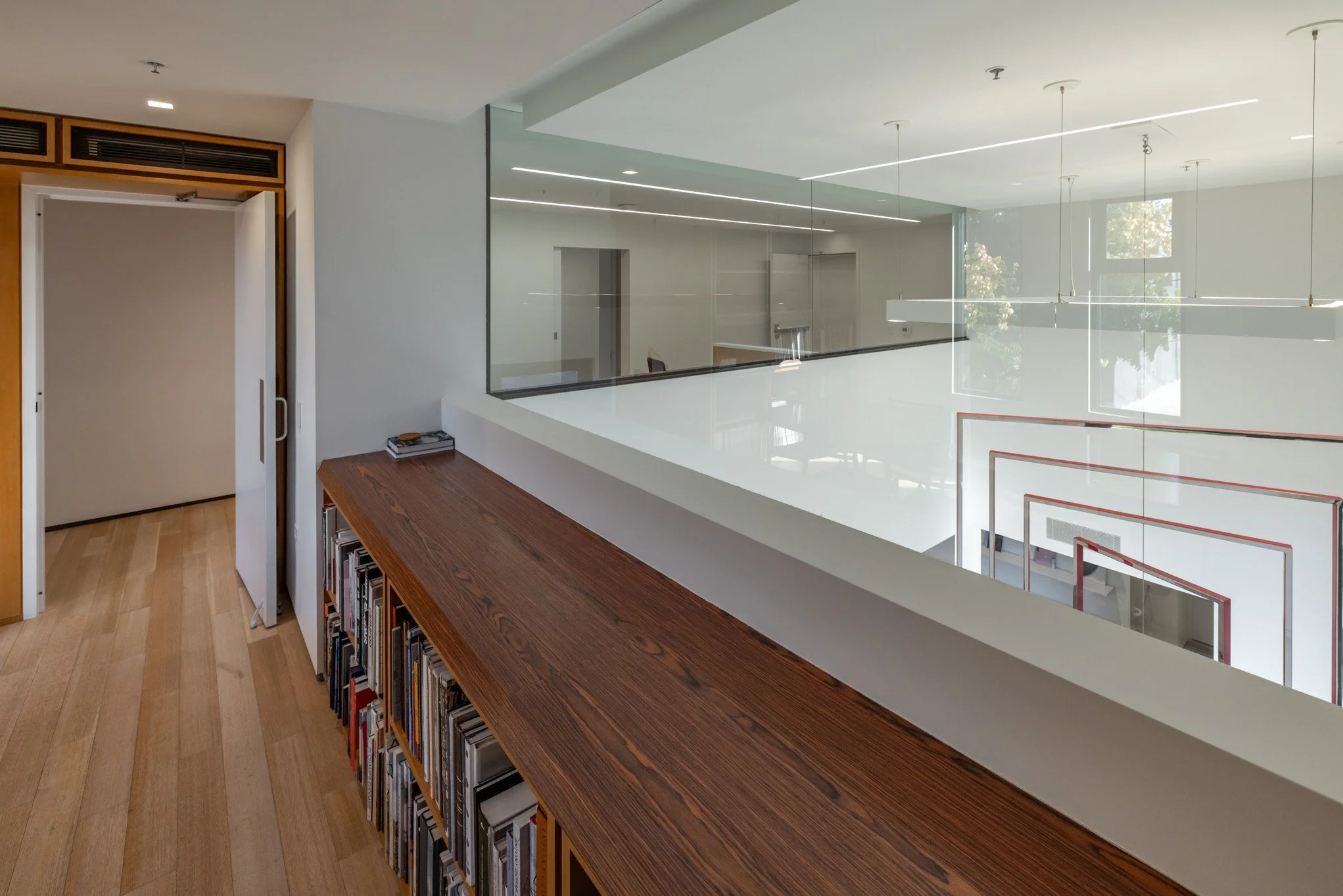 Modern interior view of a building with a wooden bookshelf, a white railing, and large windows showing trees outside.