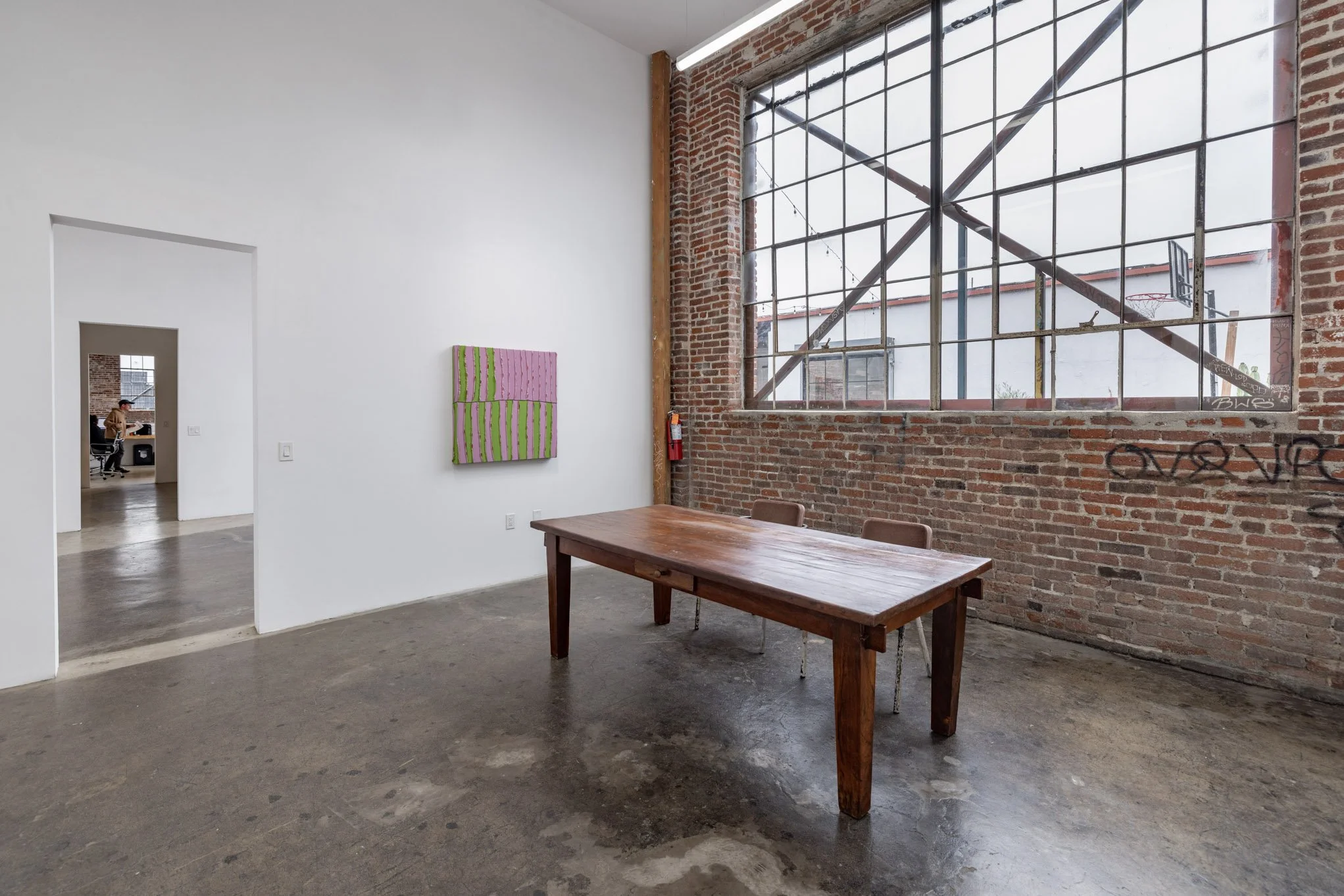 An art gallery with a wooden table and chairs, white walls, a large brick wall with a tall industrial window, and a colorful painting on the wall.
