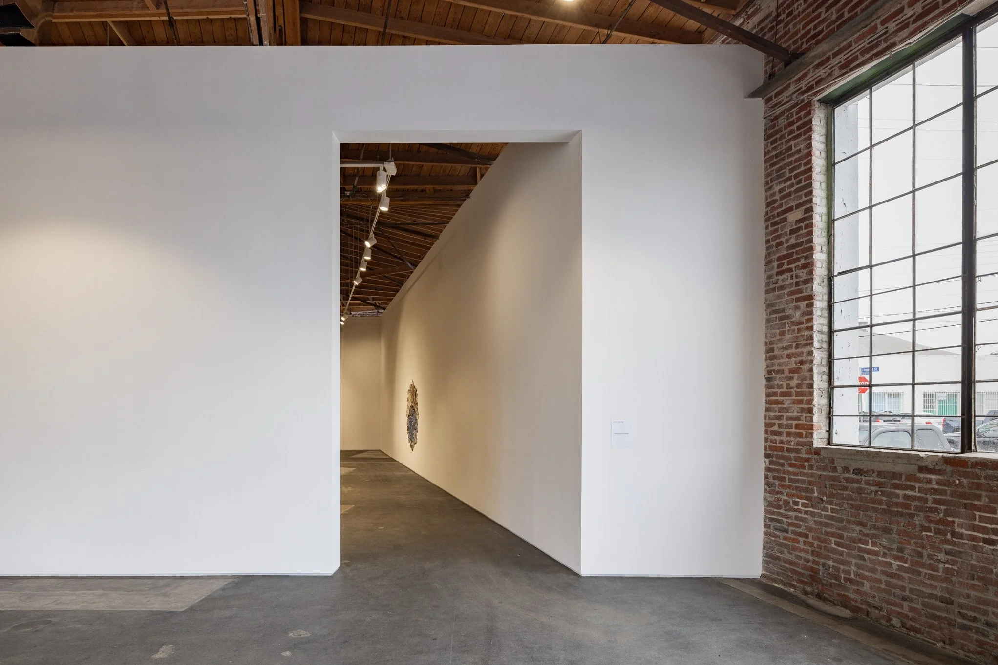 Interior of an art gallery with a white display wall, a brick wall with large windows, and wood ceiling