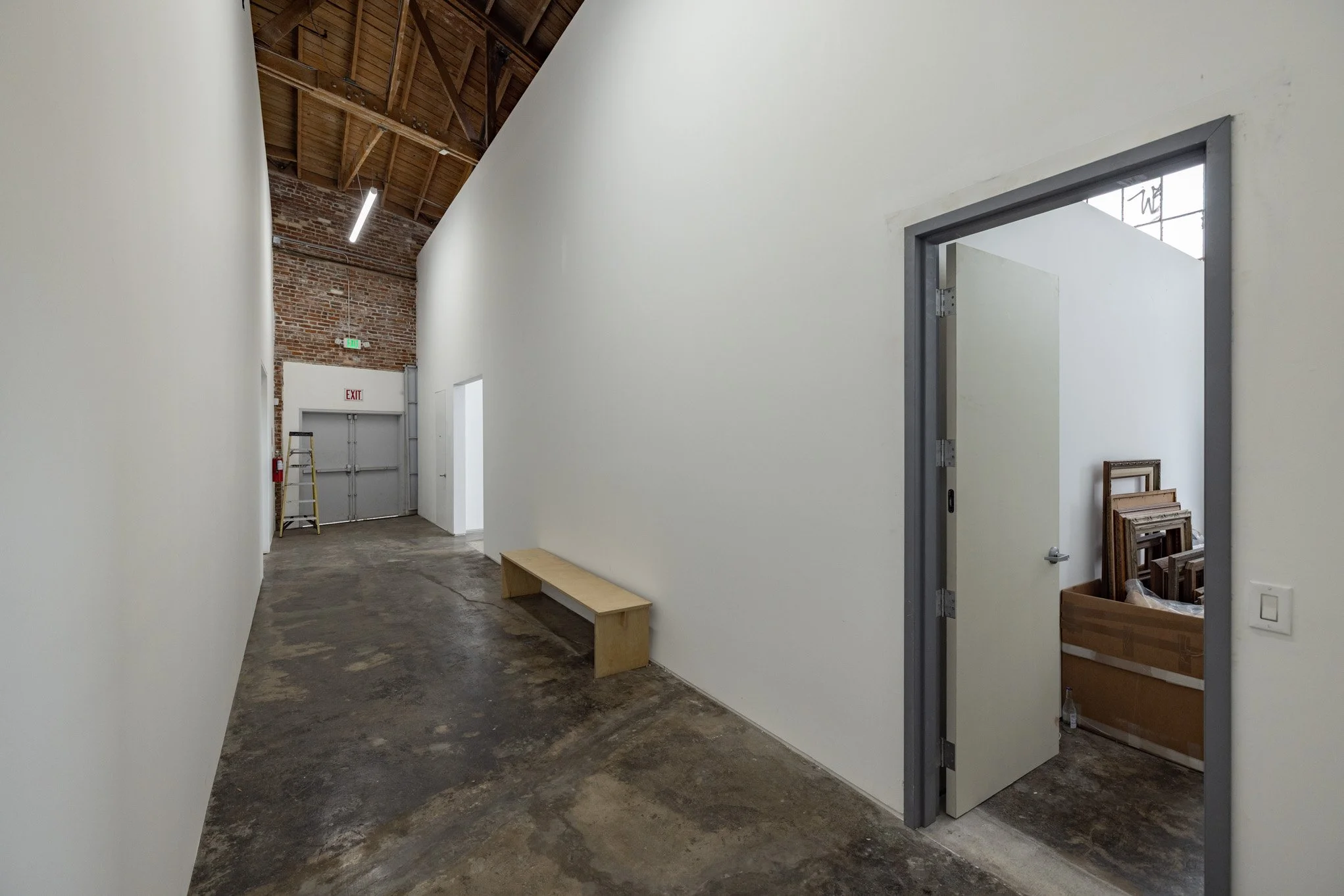 Empty hallway with white walls, wooden ceiling, concrete floor, door on the right leading to a room with picture frames inside, and glass panel above the door. Ladder, fire extinguisher, and exit door at the end of the corridor.