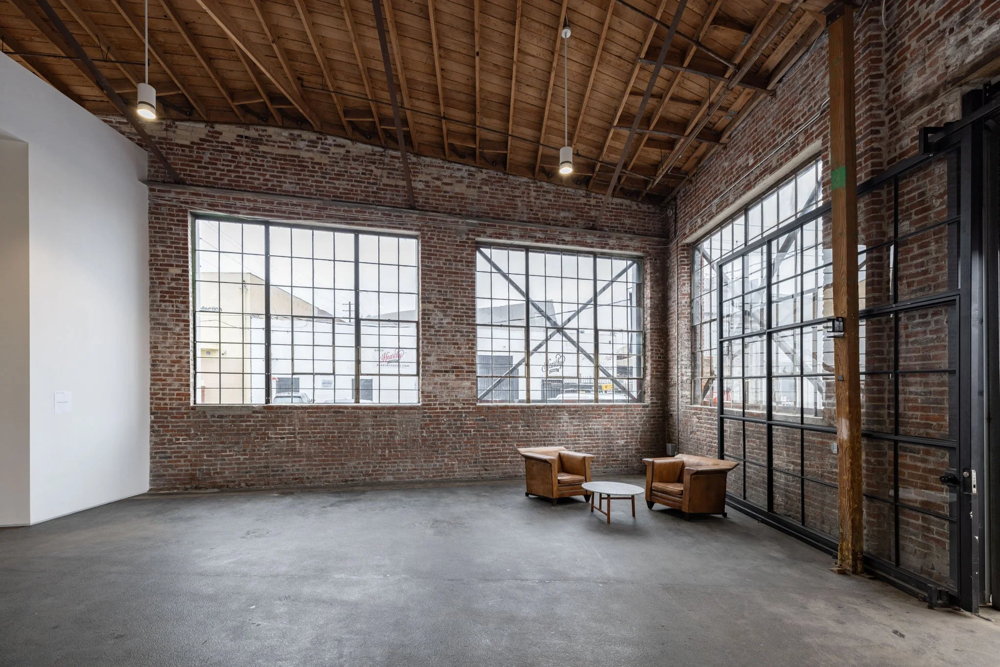 Interior of an industrial-style building with exposed brick walls, large multi-pane windows, a wooden ceiling with hanging lights, and minimal furniture including two leather chairs and a small round table.