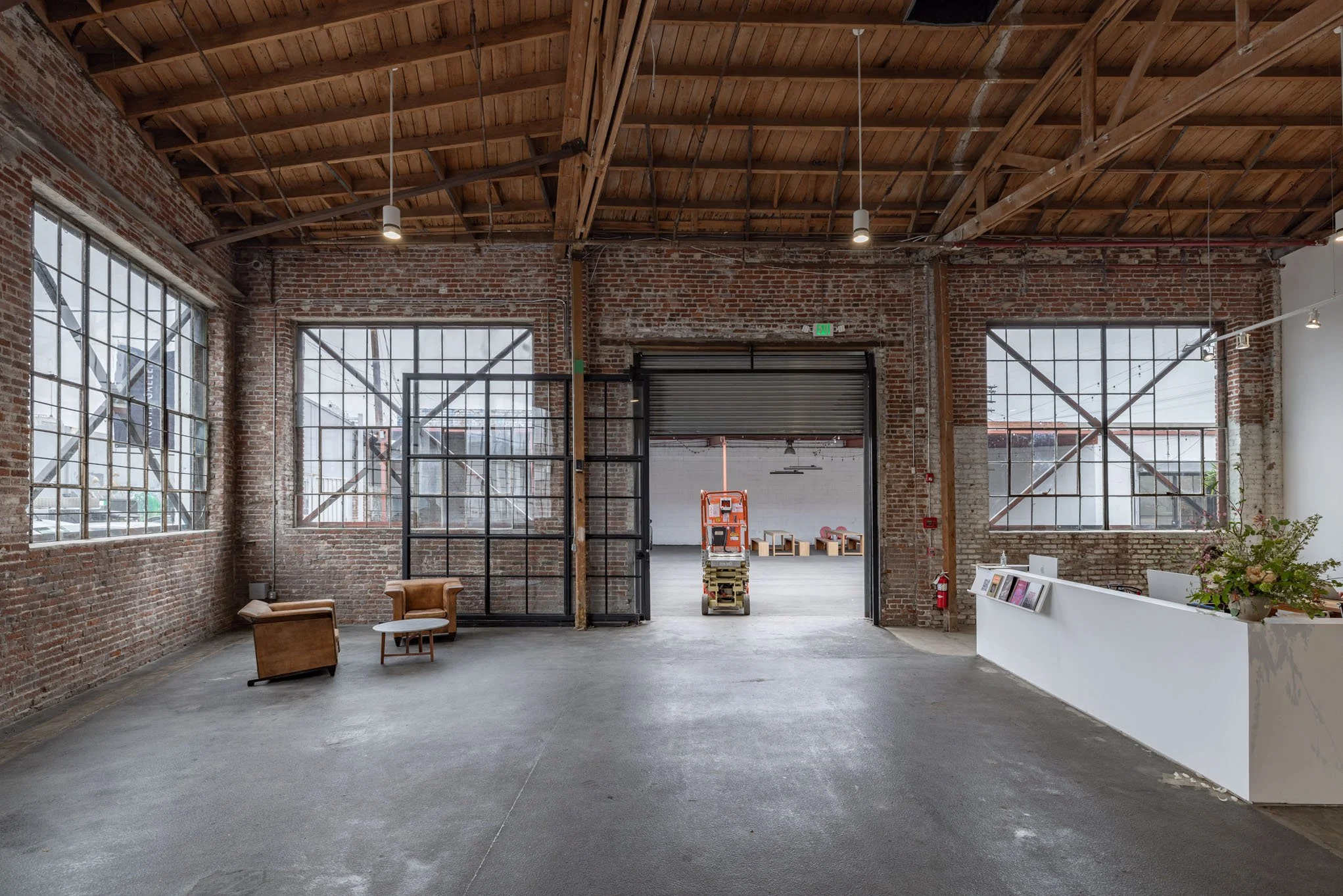 Industrial-style interior with exposed brick walls, large windows, wooden ceiling, leather armchairs, reception desk, and a lift door leading to a spacious open area.