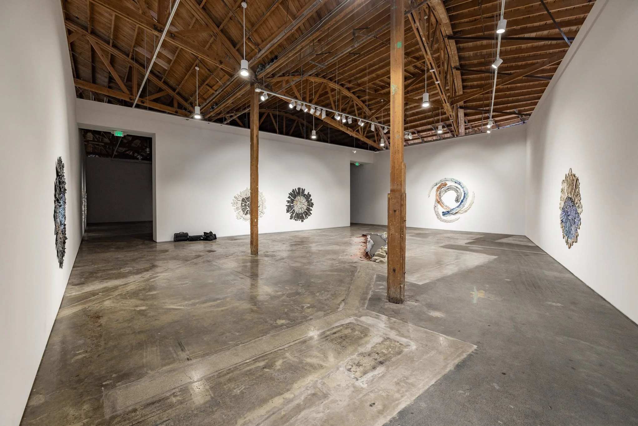 An art gallery with white walls, wooden ceiling beams, and artwork hanging on the walls, featuring abstract and textured designs.