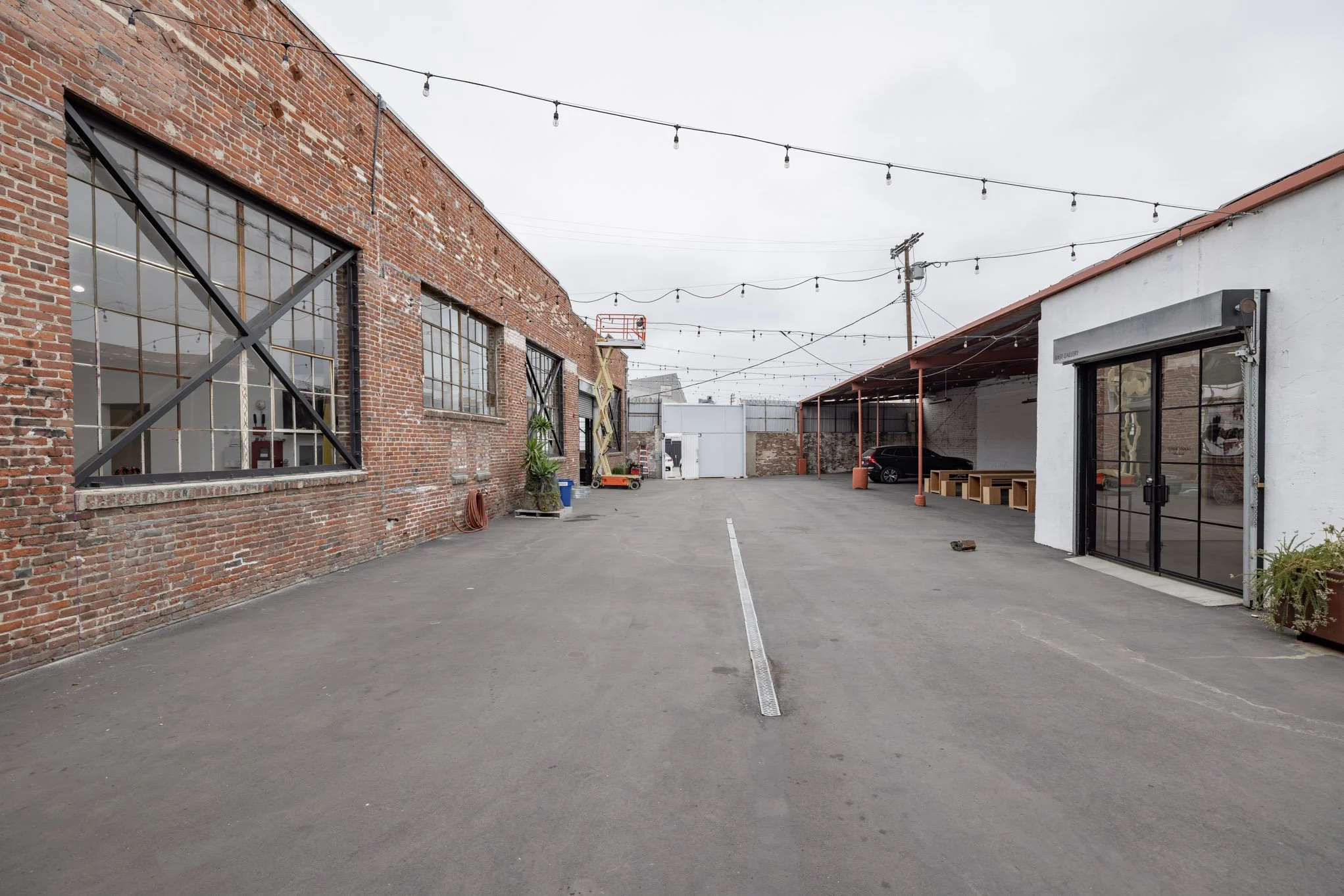 Empty urban courtyard with brick and white buildings, string lights overhead, and parked cars.