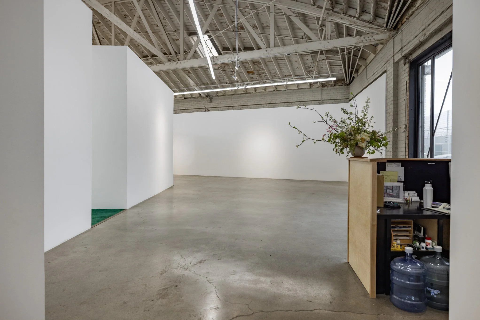 Empty art gallery with white walls, a concrete floor, and an industrial ceiling with exposed beams and fluorescent lights. There is a small reception desk with a flower arrangement and water bottles in the corner near a large window.