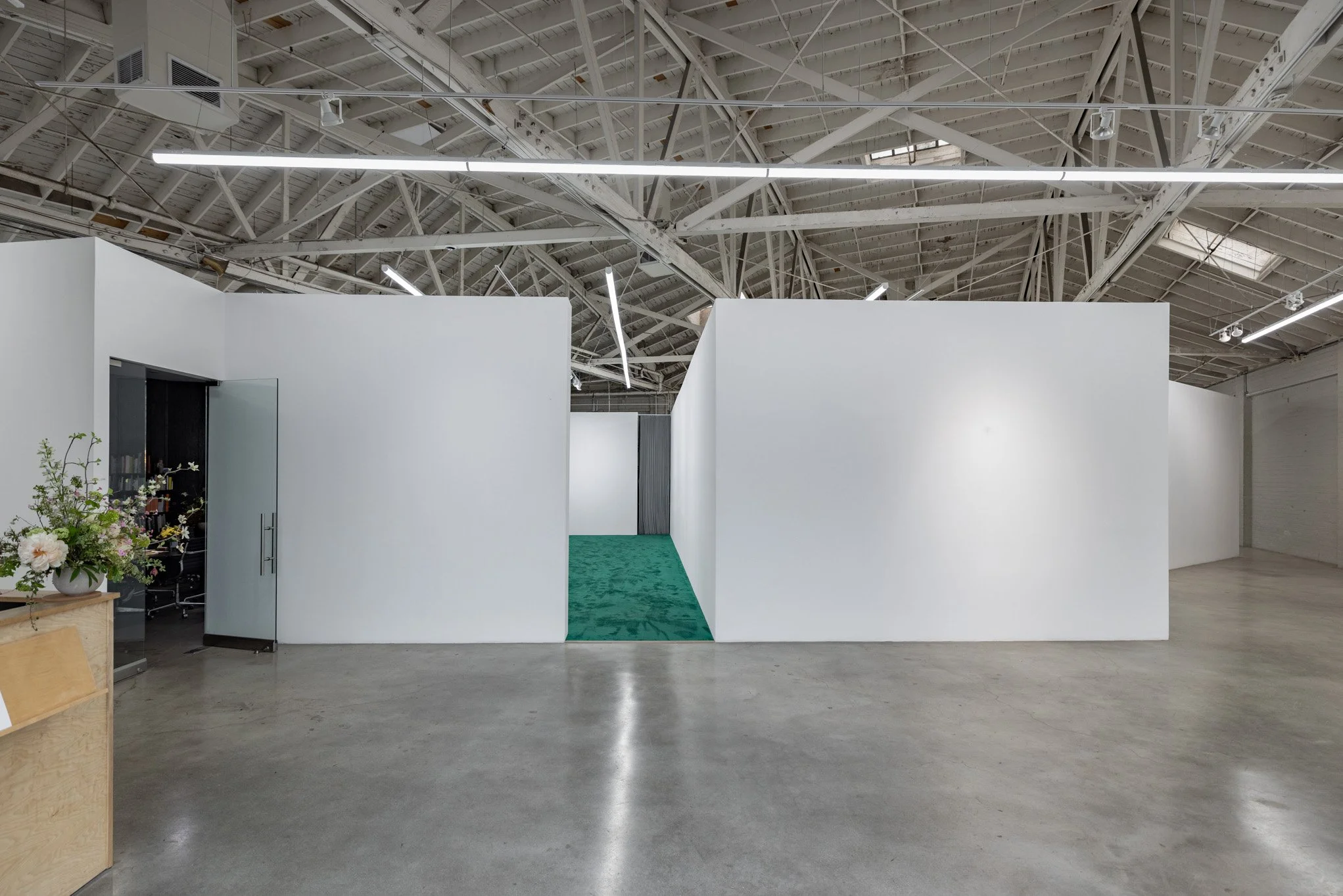 Empty art gallery with white walls, a green carpeted walkway, and industrial-style ceiling with exposed beams and fluorescent lighting.