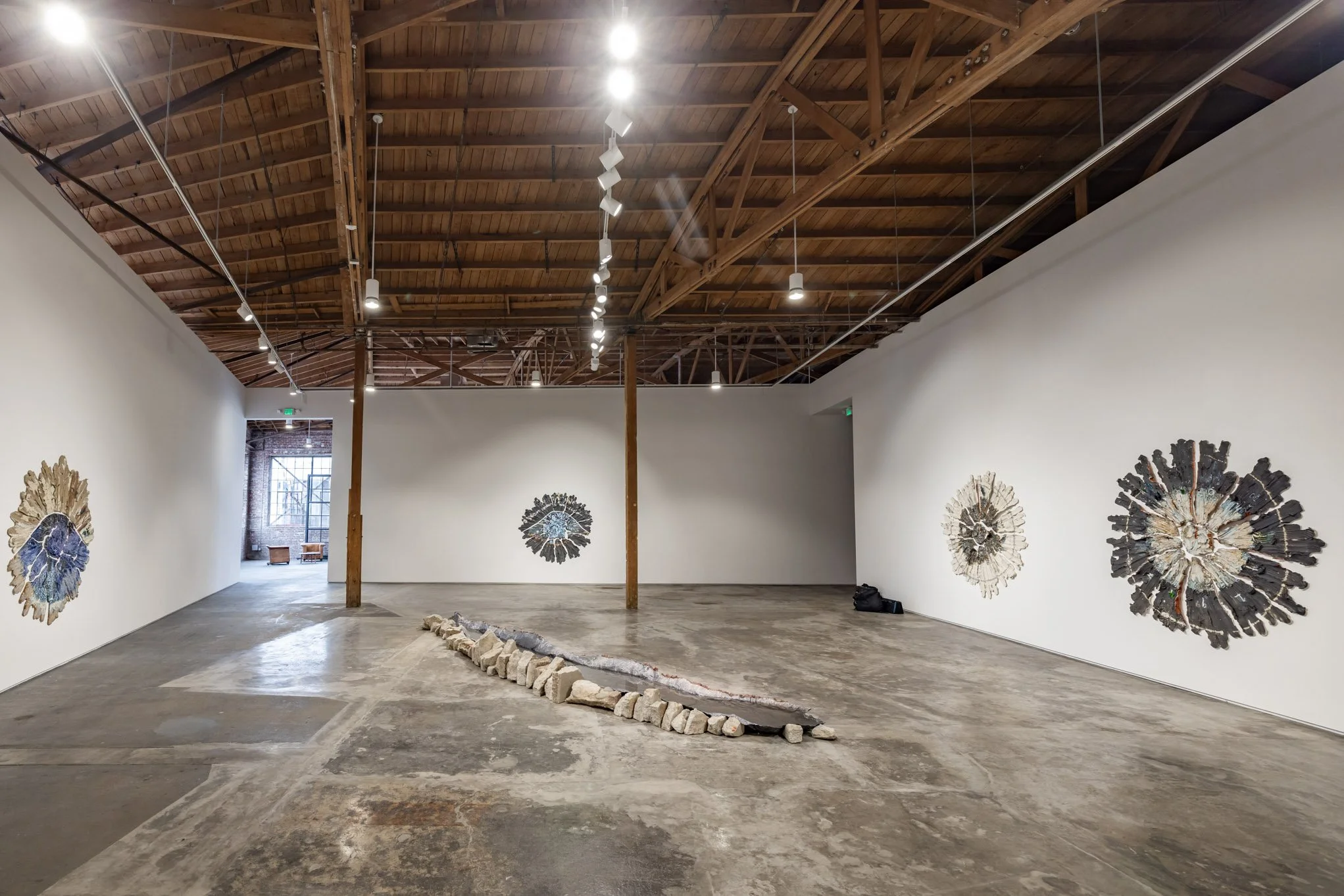 An art gallery with white walls, a wooden ceiling, and concrete floors. Several abstract circular artworks are displayed on the walls, and a long, curved sculpture made of rocks is positioned on the floor in the center.