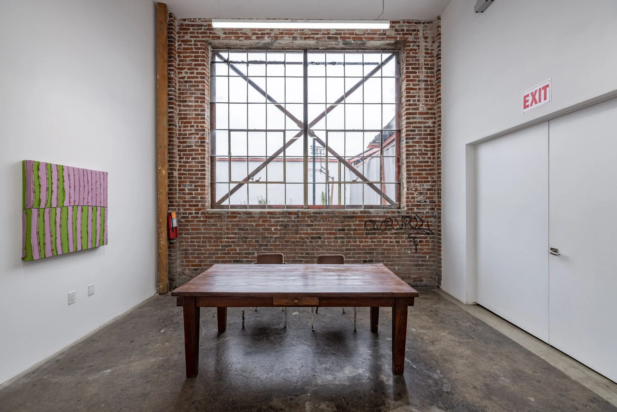 An art gallery with a brick wall and a large window with security bars. A wooden table and two chairs are in the center. There's graffiti on the brick wall, a pink and green striped artwork on the white wall, an EXIT sign, and a fire extinguisher.