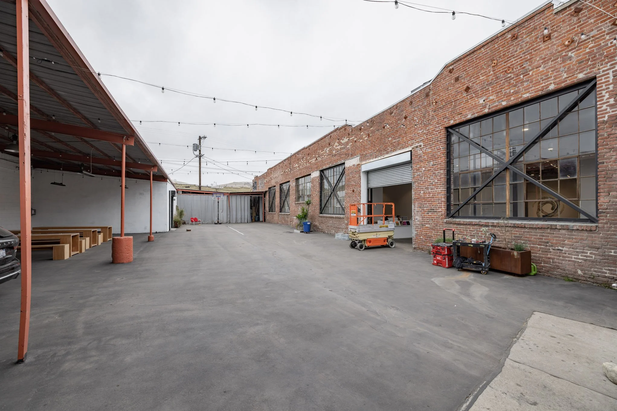 An industrial brick building with large windows and a roller door in an outdoor parking lot. The parking lot has string lights overhead and some scattered equipment and lounge chairs.