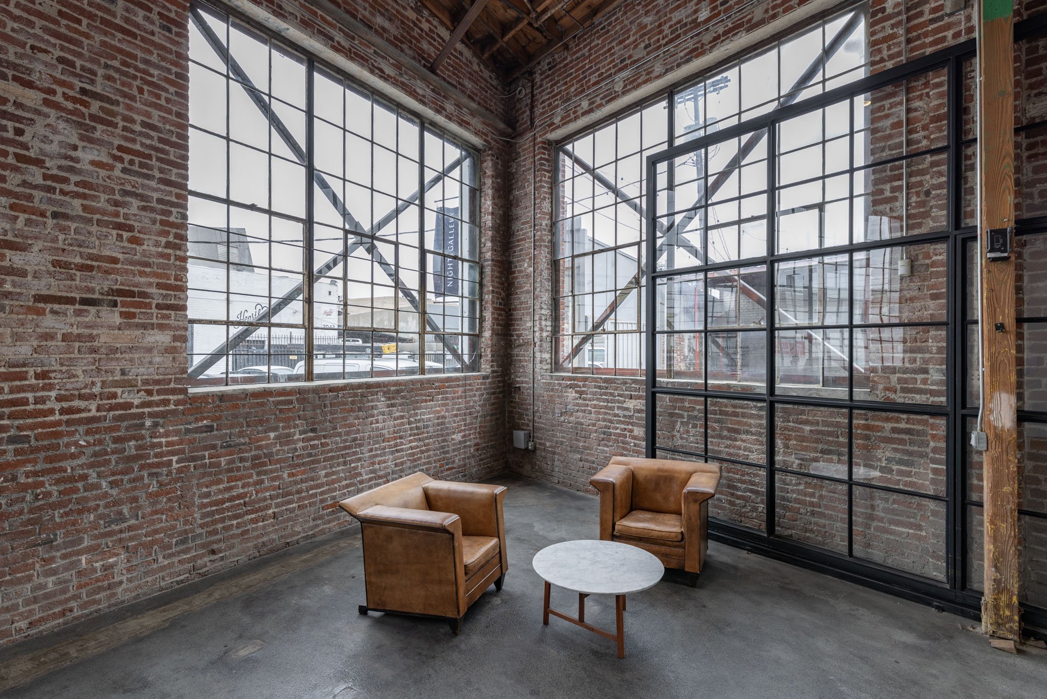 Interior of an industrial-style loft with exposed brick walls, large grid windows, two brown leather armchairs, and a small round marble-topped coffee table.
