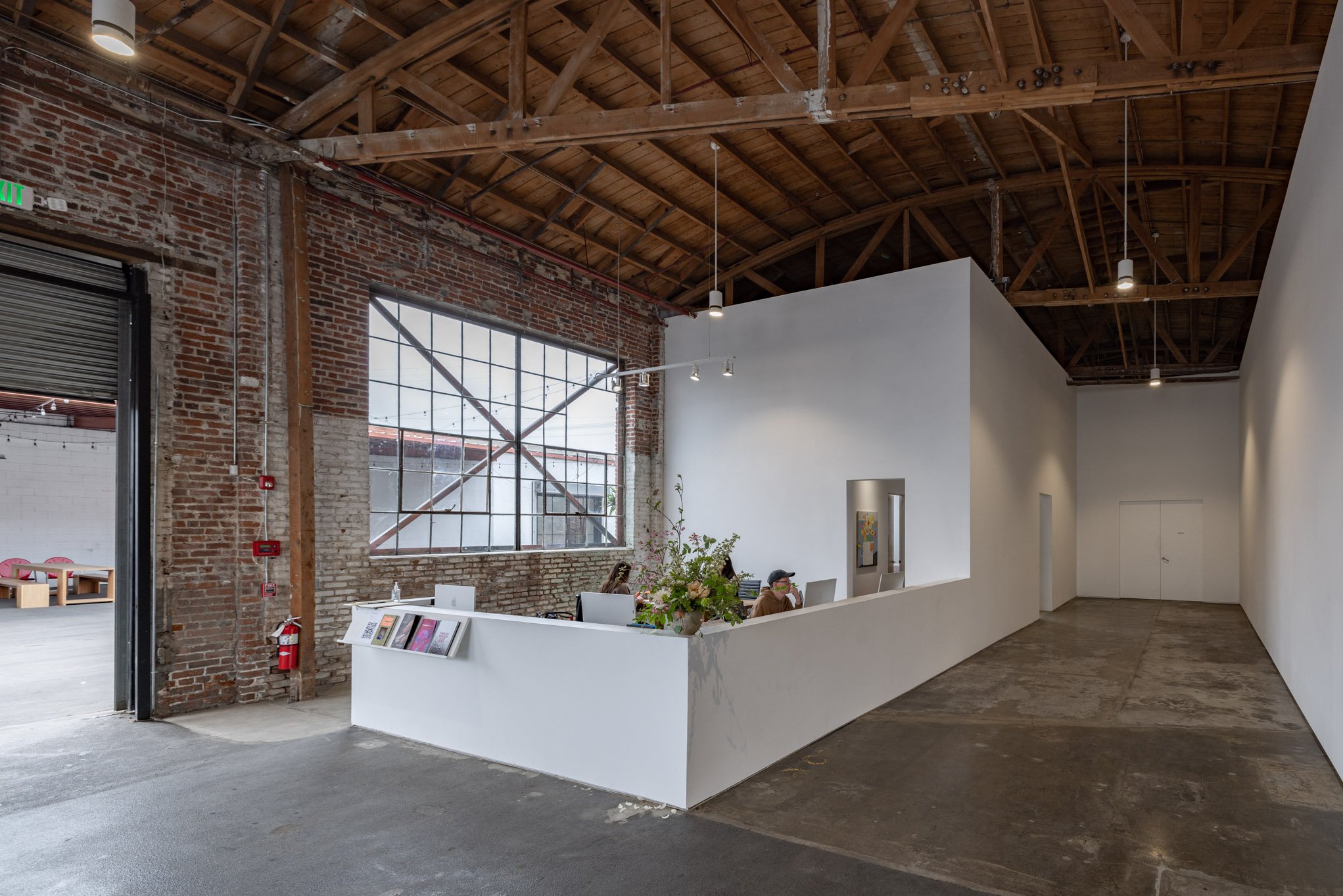 An industrial-style interior space with exposed brick walls, large grid windows, a white reception desk with people working behind it, and a high wooden ceiling with pendant lights.