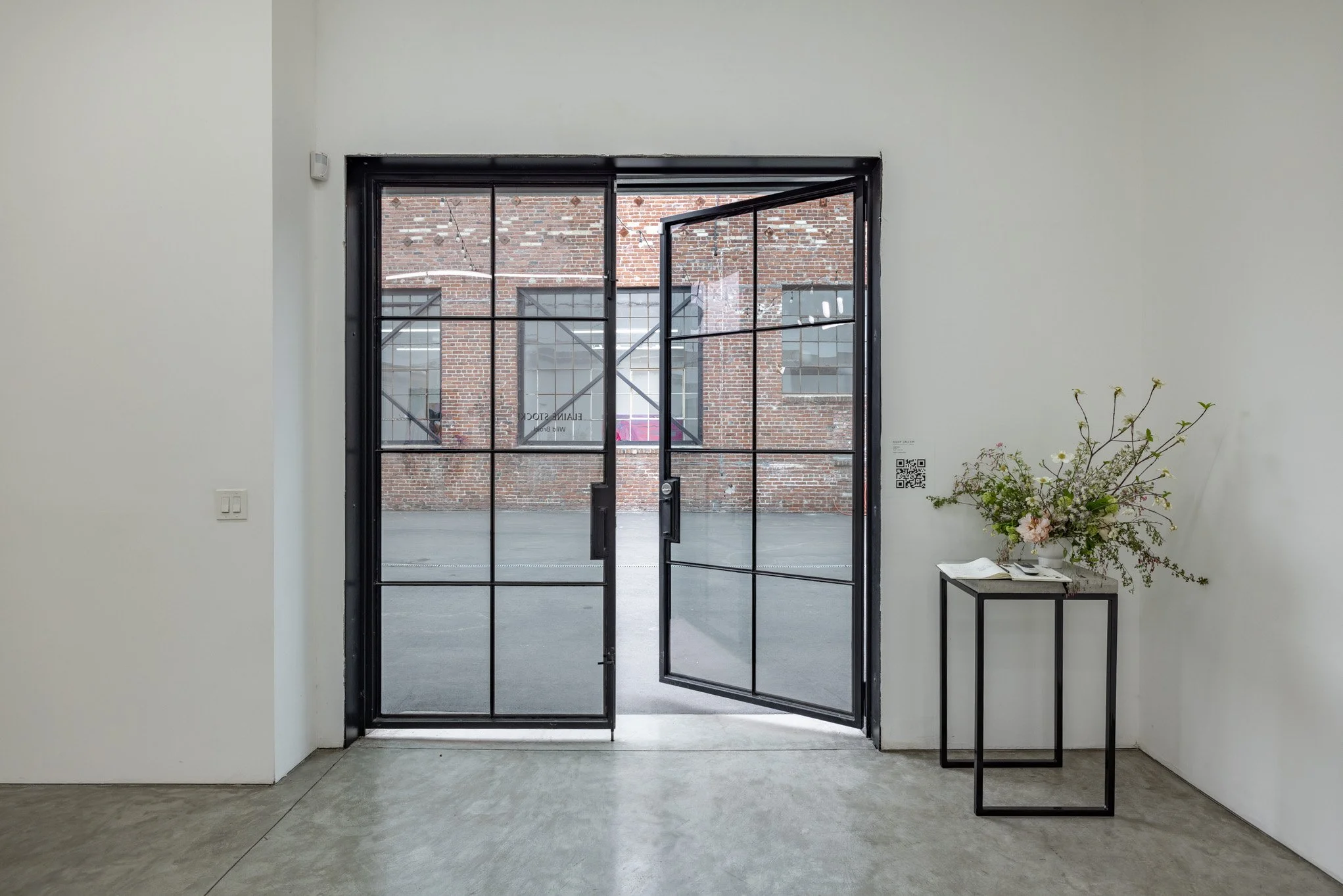 Interior view of a minimalistic room with glass double doors opening to an outdoor brick wall. A black metal side table with a large floral arrangement and some papers is on the right.