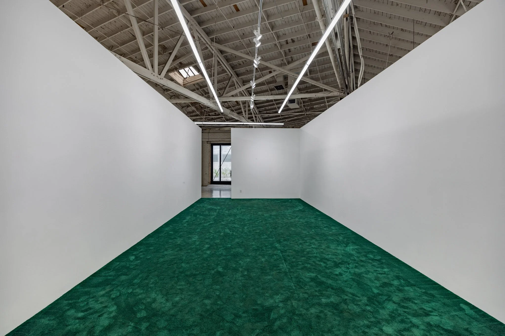 An empty art gallery with white walls, green carpet, exposed ceiling beams, and linear hanging lights.