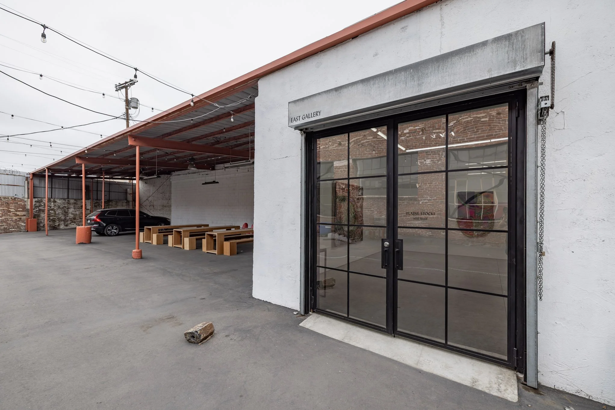 Exterior of an art gallery called East Gallery, with a glass door and benches outside. There is a covered parking space with a black car and string lights overhead.
