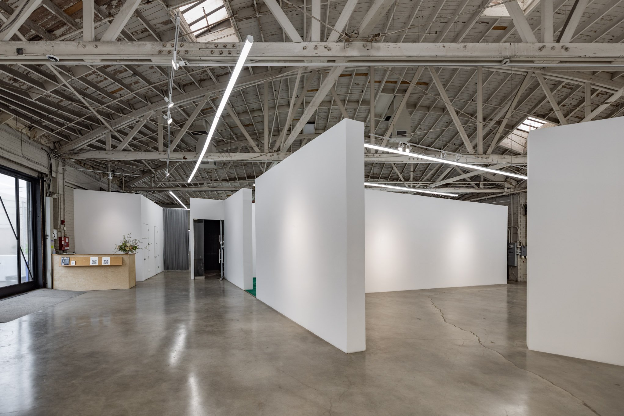 An art gallery with white walls and a concrete floor inside an industrial-style building with exposed beams and skylights.