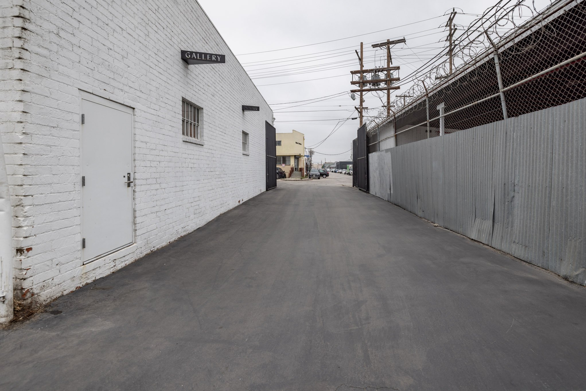 A narrow alleyway with a white brick building on the left labeled 'GALLERY' and a corrugated metal fence with barbed wire on the right, leading to a parking lot with cars and power lines overhead.