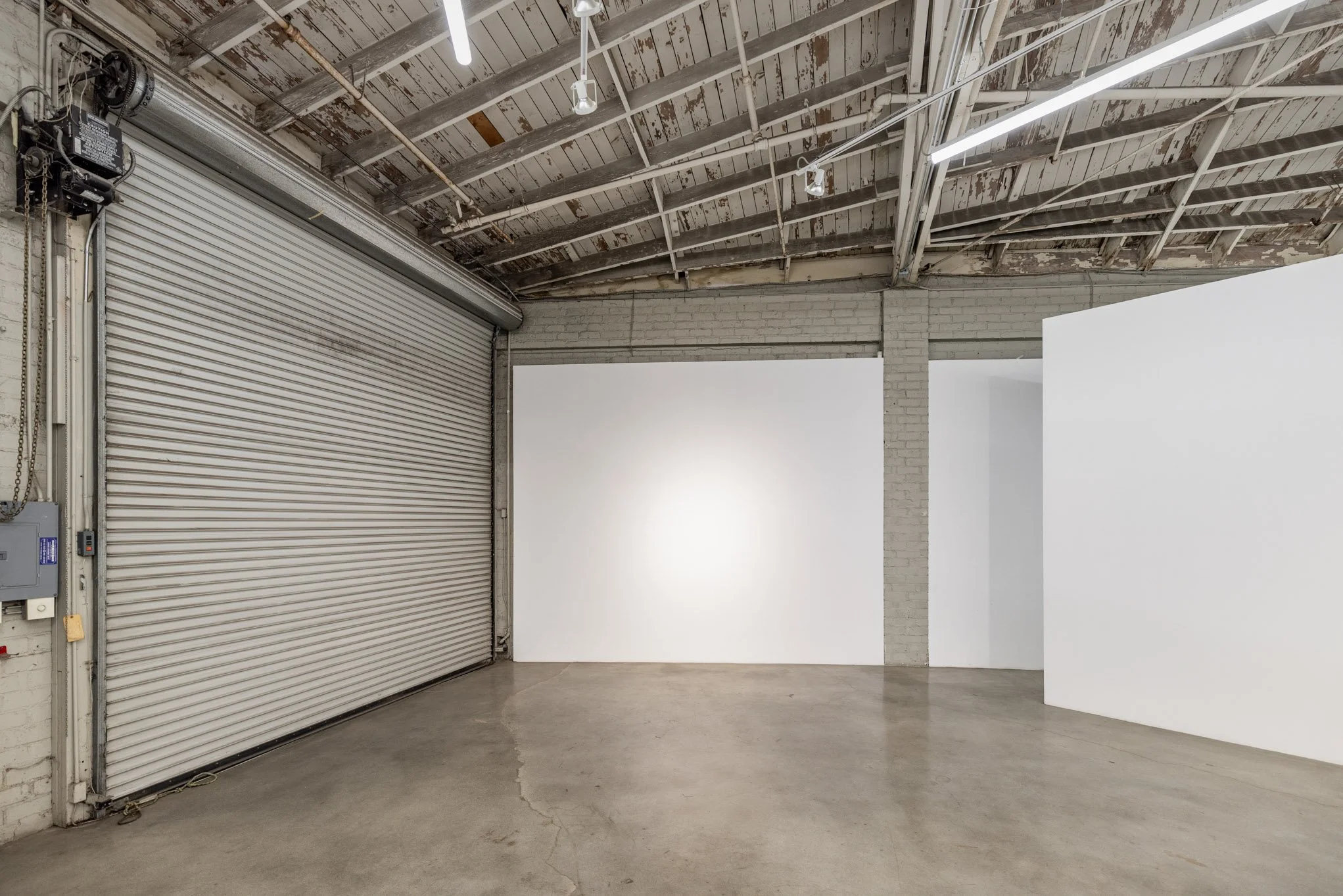 Empty interior space with concrete floor, white walls, and an industrial ceiling with exposed beams and lighting. There is a gray rolling metal door on the left side.