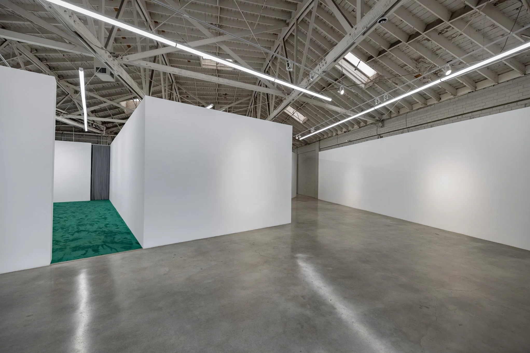 Empty art gallery with white walls, concrete floor, and visible ceiling structural beams with fluorescent lighting.