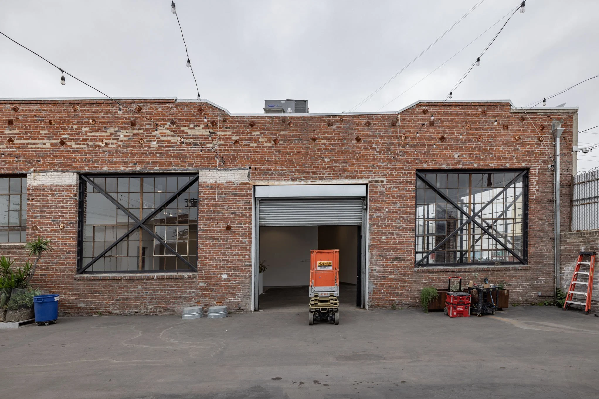 The image shows the front of a brick building with large industrial-style windows protected by metal cross braces, and a partially open rolling garage door in the center. There is a small lift platform and various tools and equipment outside. String 