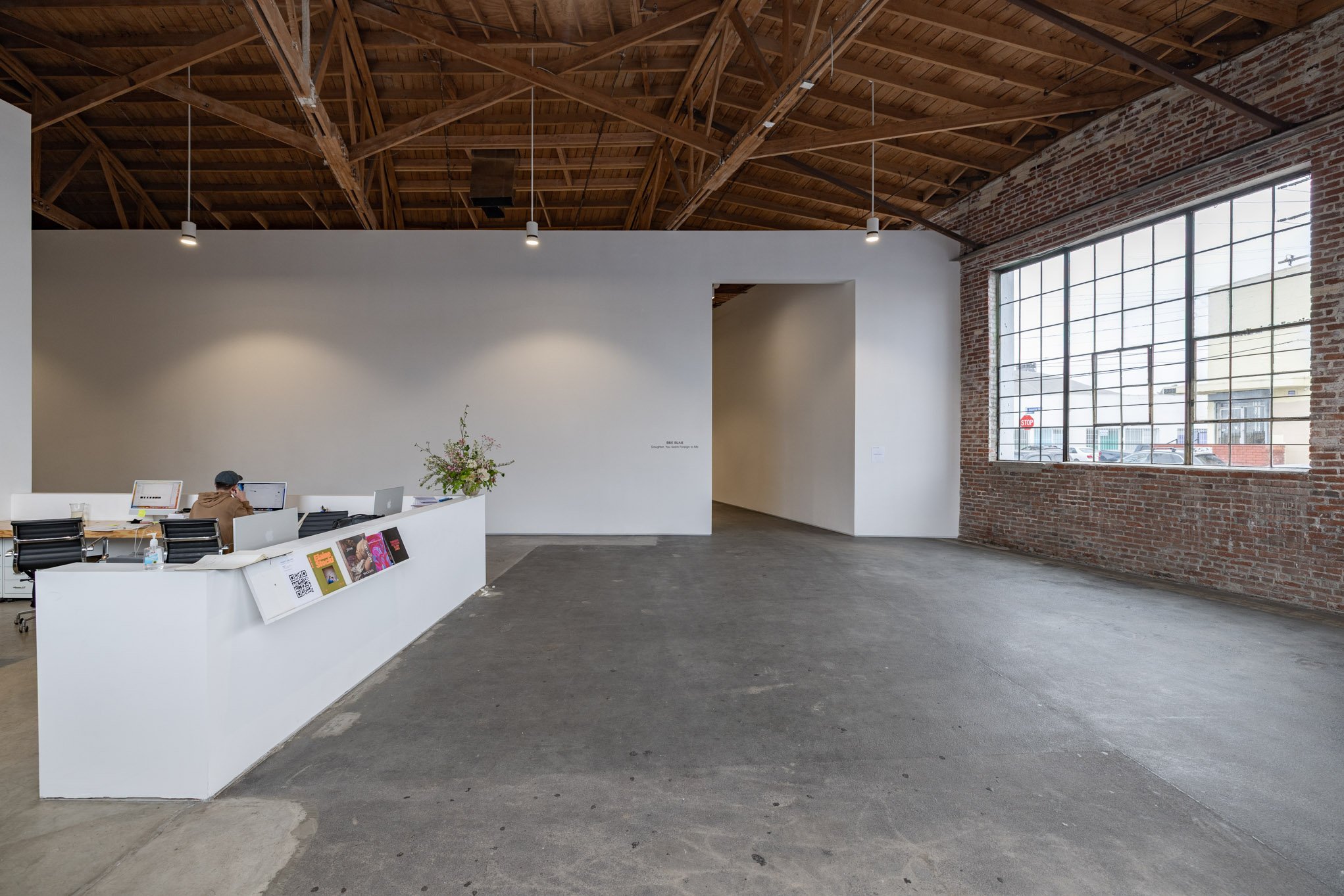 A spacious, industrial-style office with a high wooden ceiling, exposed brick wall, large metal-framed window, and a white reception desk with two people working behind it.