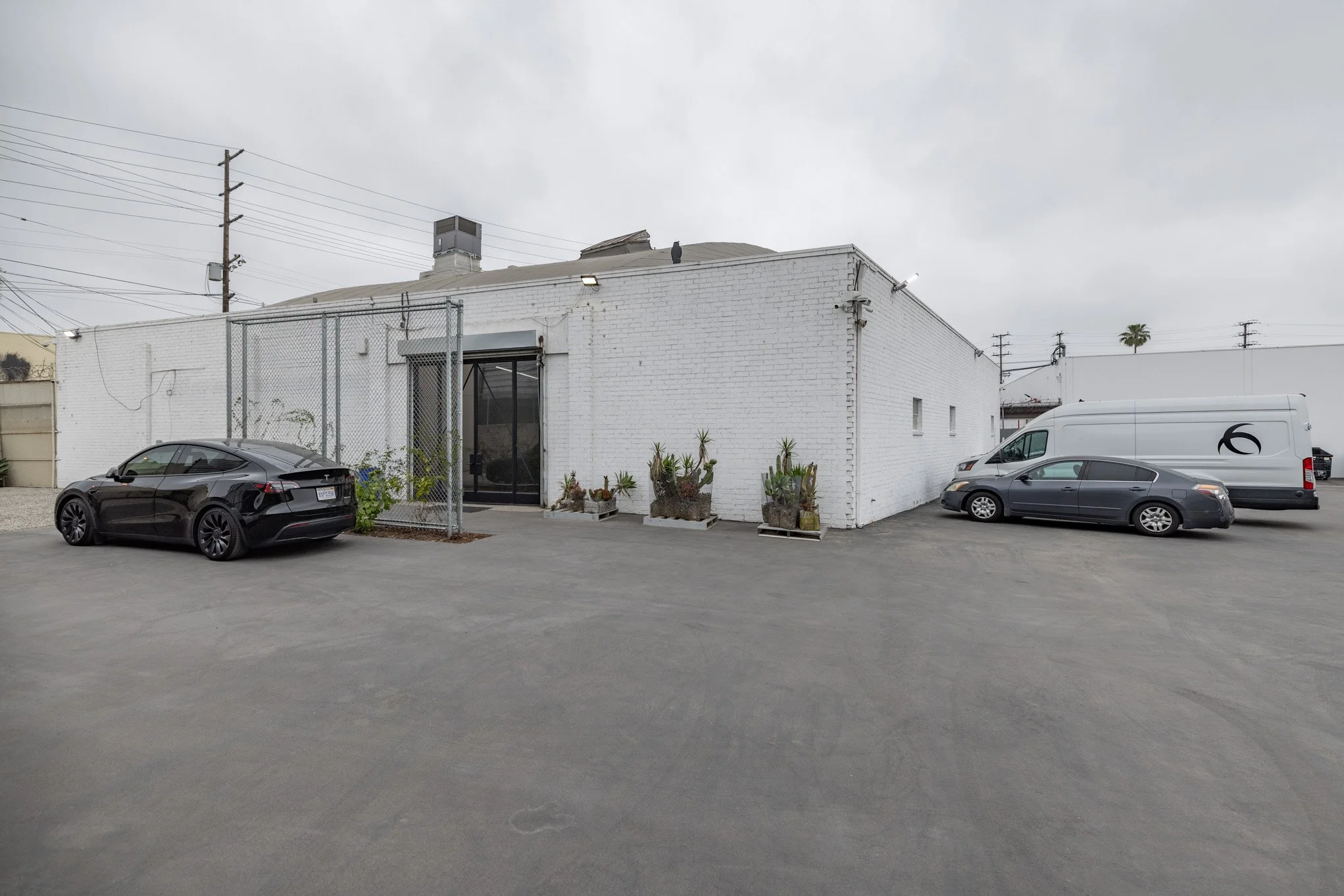 A parking lot with three parked vehicles in front of a white brick building, some plants in concrete containers, and power lines overhead under a cloudy sky.