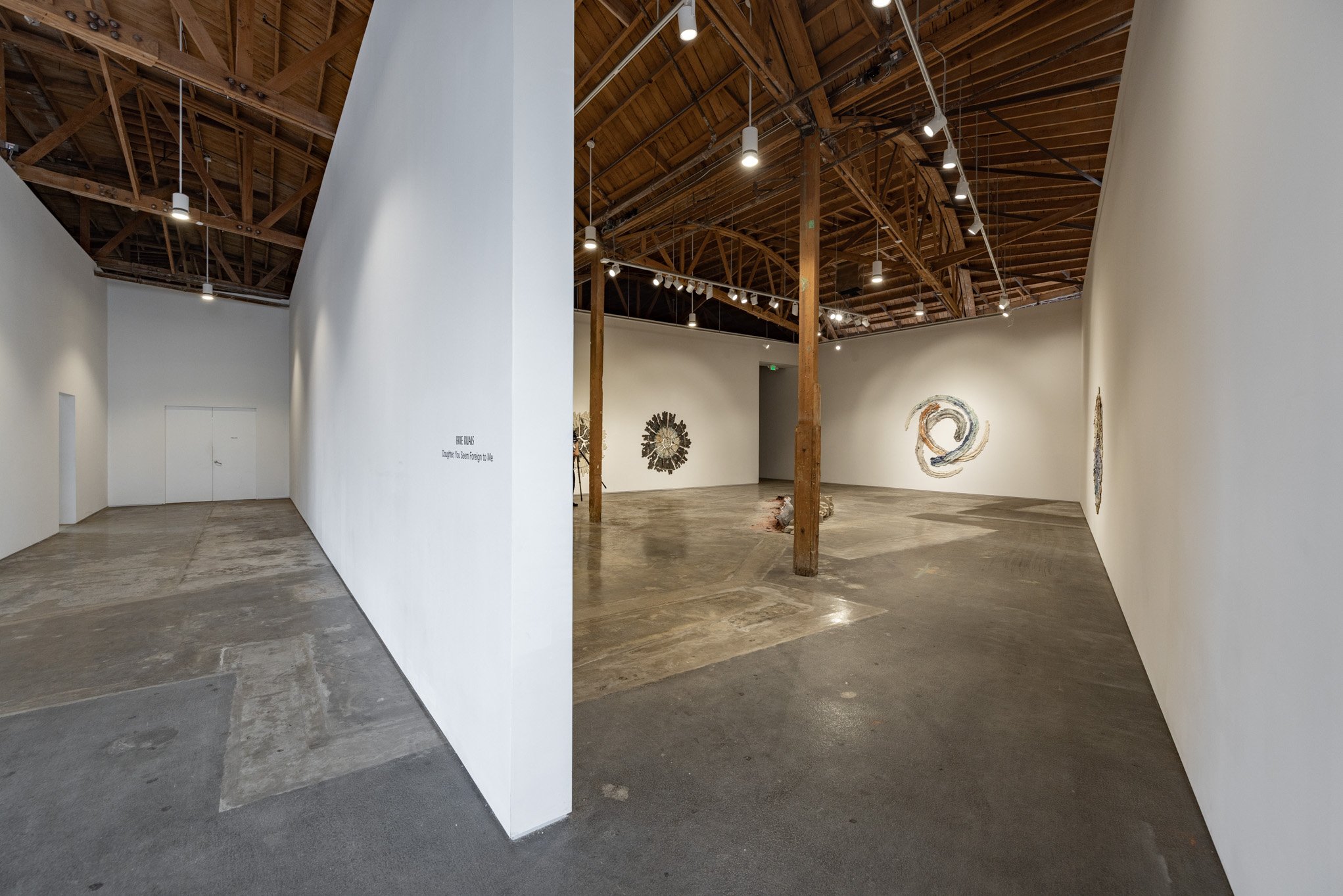Interior view of an art gallery with white walls, wooden ceiling, and concrete floor, displaying various abstract art pieces on the walls.