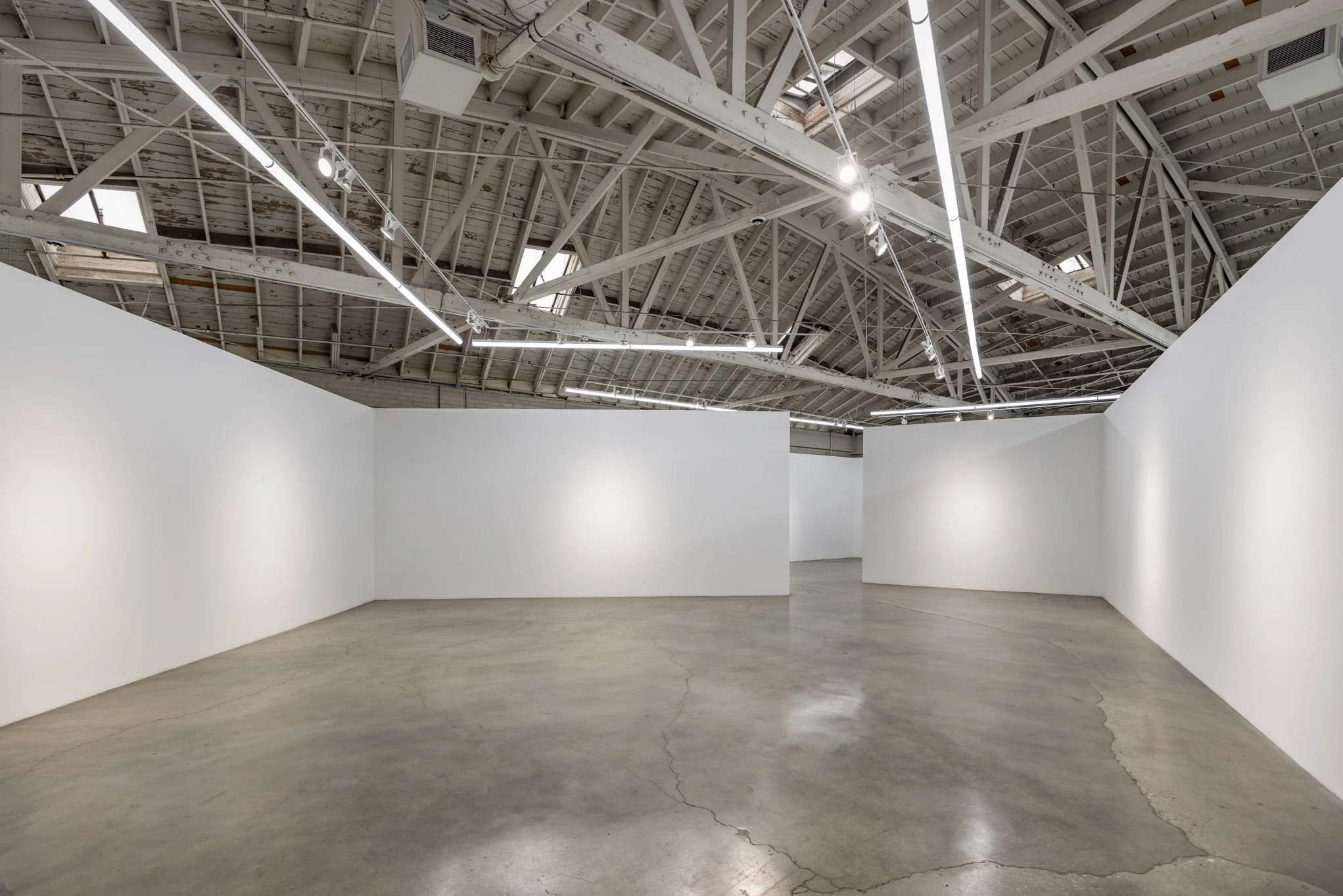 Empty art gallery with white walls, concrete floor, and exposed ceiling with pipes and lighting fixtures.