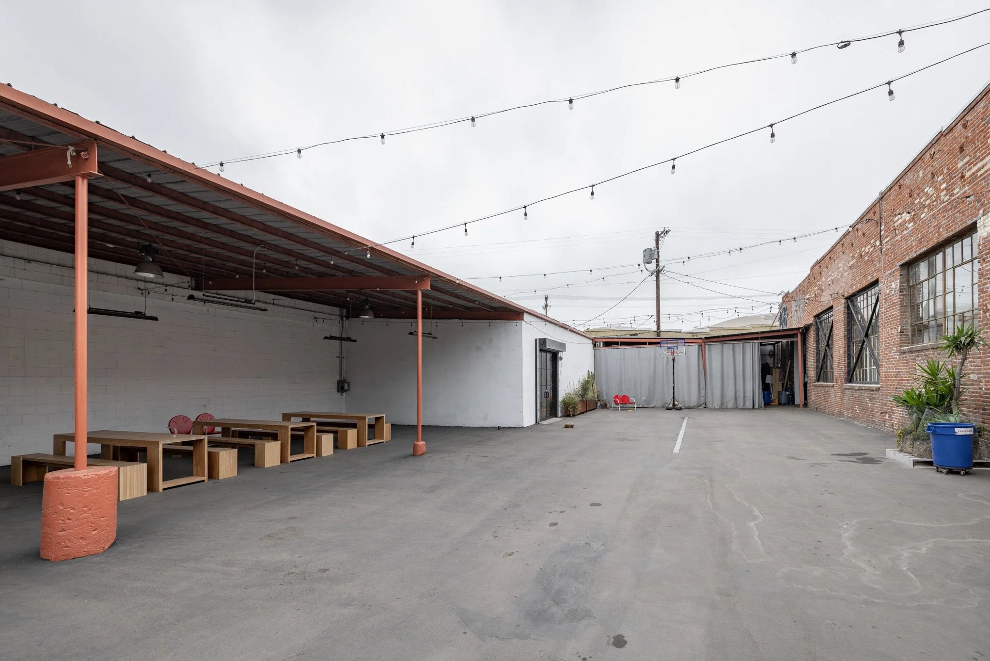Empty outdoor patio area with concrete ground, brick and white walls, string lights overhead, benches under a covered patio, a basketball hoop, and a few chairs near the back.