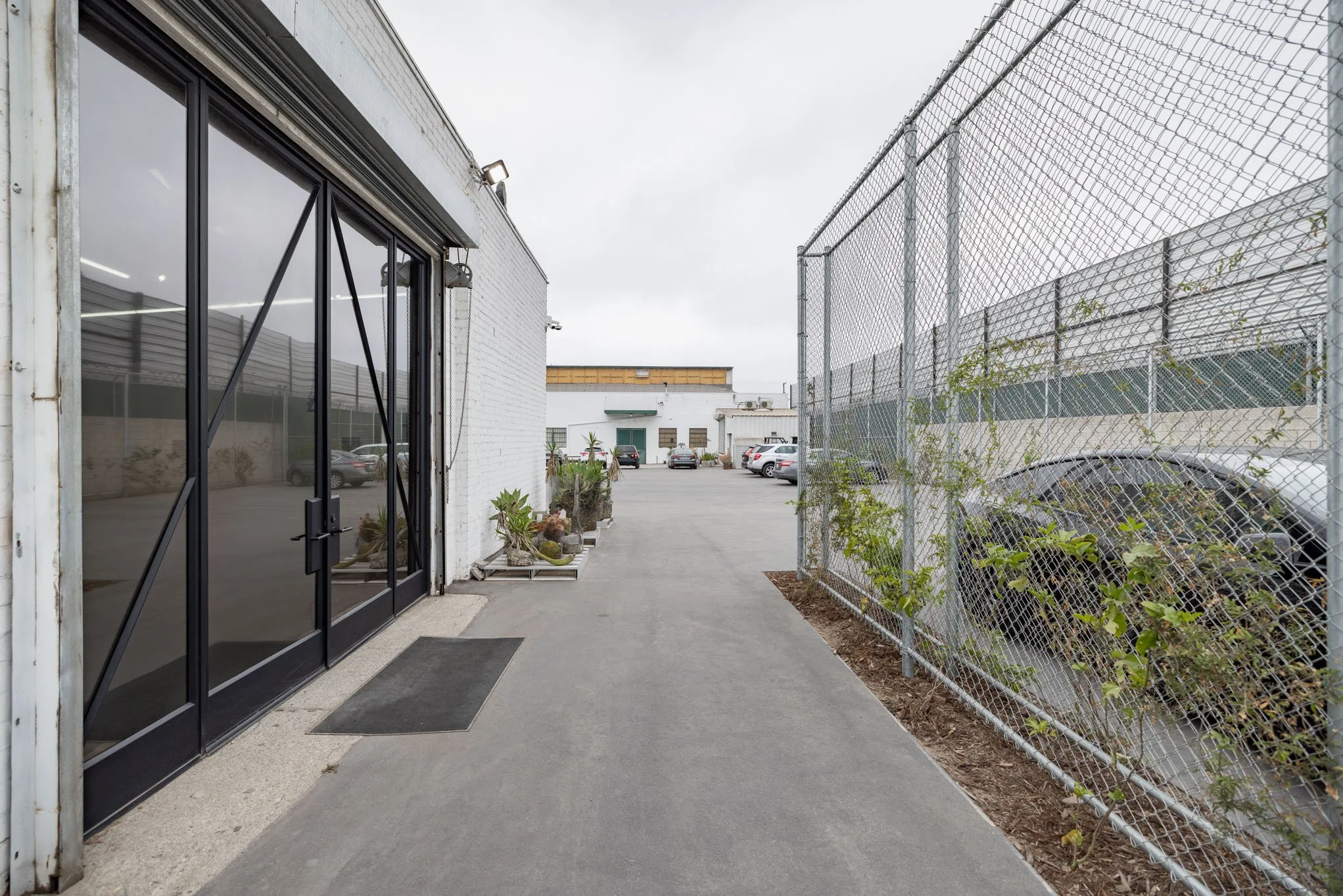 Exterior view of an industrial or commercial building with glass doors, potted plants, a fenced parking lot, and parked cars under an overcast sky.