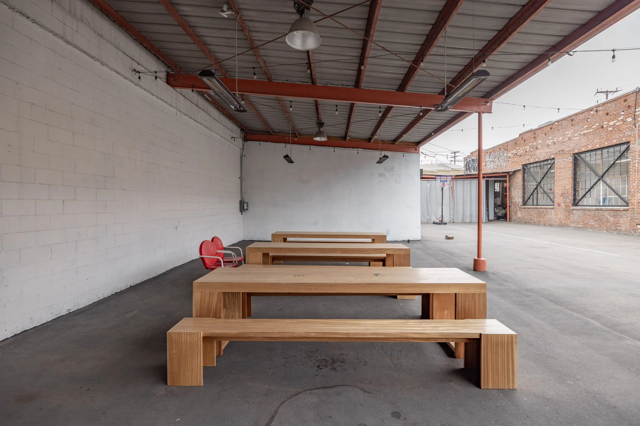 Empty outdoor patio with wooden tables and benches under a metal roof, brick and white wall exterior, string lights, and red chairs at the back.