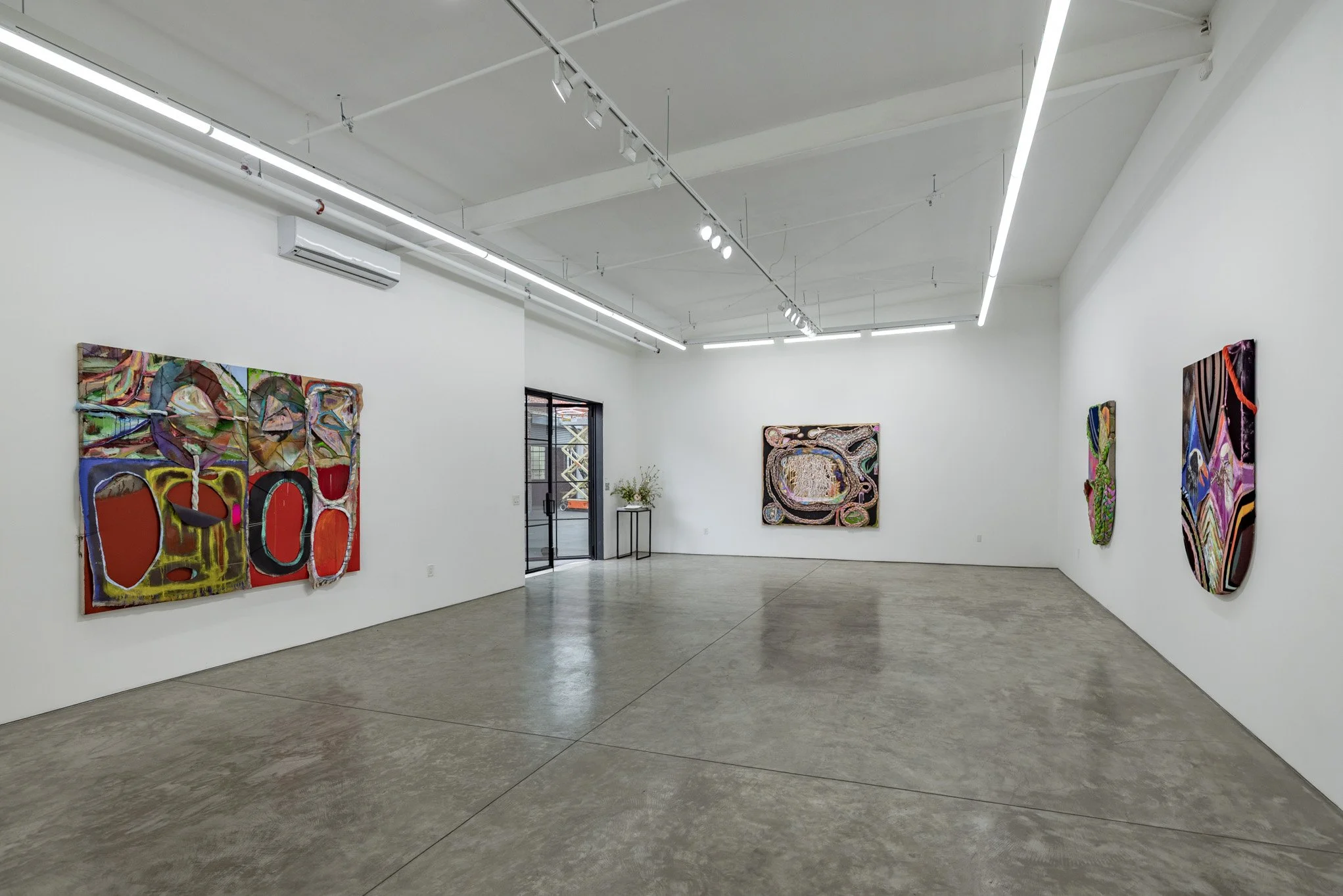 An art gallery with white walls displaying several colorful abstract paintings. The gallery has polished concrete floors and modern lighting fixtures.