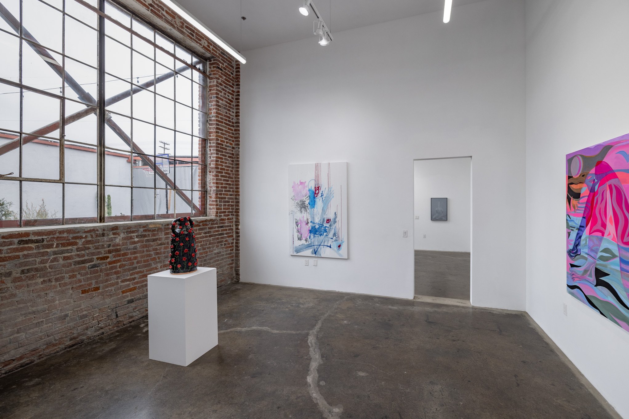 Art gallery with white walls, contemporary paintings, a sculpture with a black base and red accents, large window with metal grid, concrete floor.