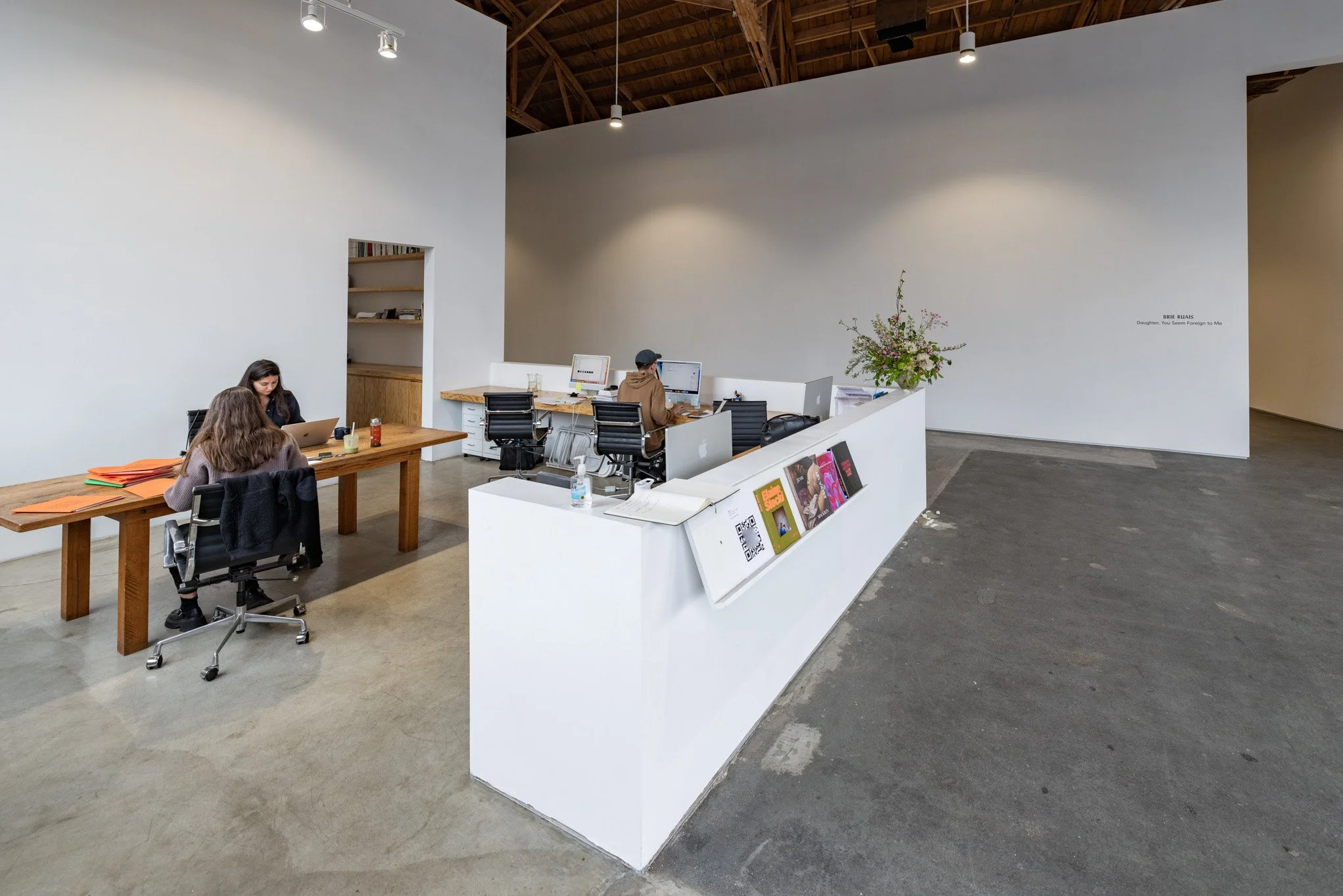 A modern, minimalistic office space with three employees working at separate desks. The office has white walls, exposed wooden ceiling beams, and polished concrete floors. One employee is working on a laptop at a wooden table, while two others are at