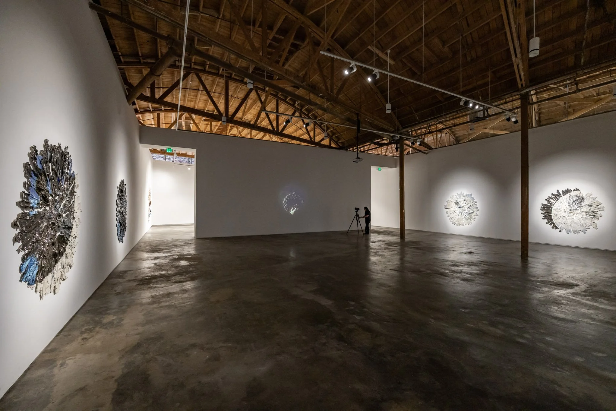 An art gallery with white walls and a wooden ceiling displaying metallic art pieces that resemble abstract flowers or explosions. A person is filming with a tripod in the distance, with projected images on the wall.