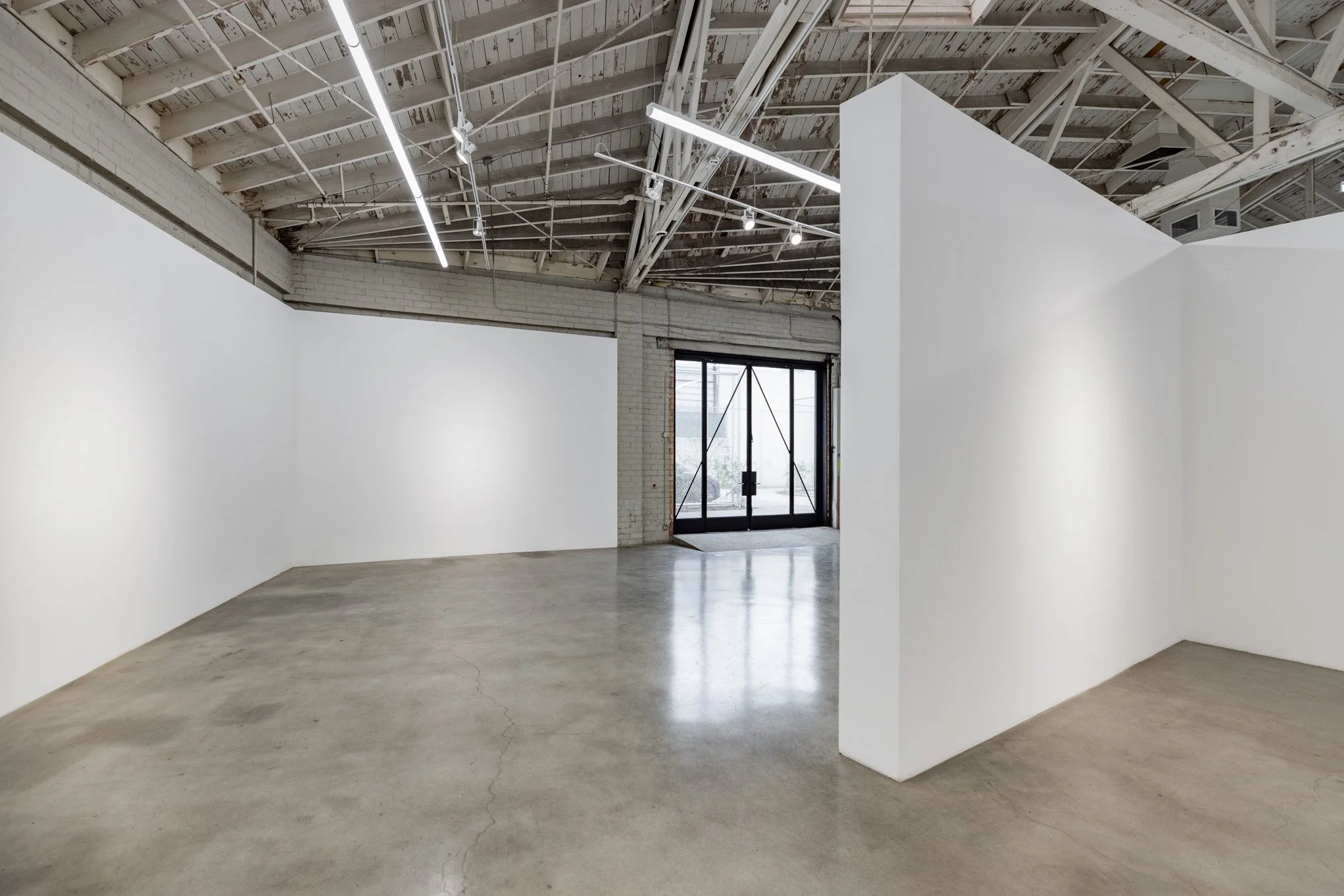 An empty art gallery or studio with white walls, a concrete floor, and an industrial ceiling with exposed beams and lighting. There is a door with a glass panel at the back.