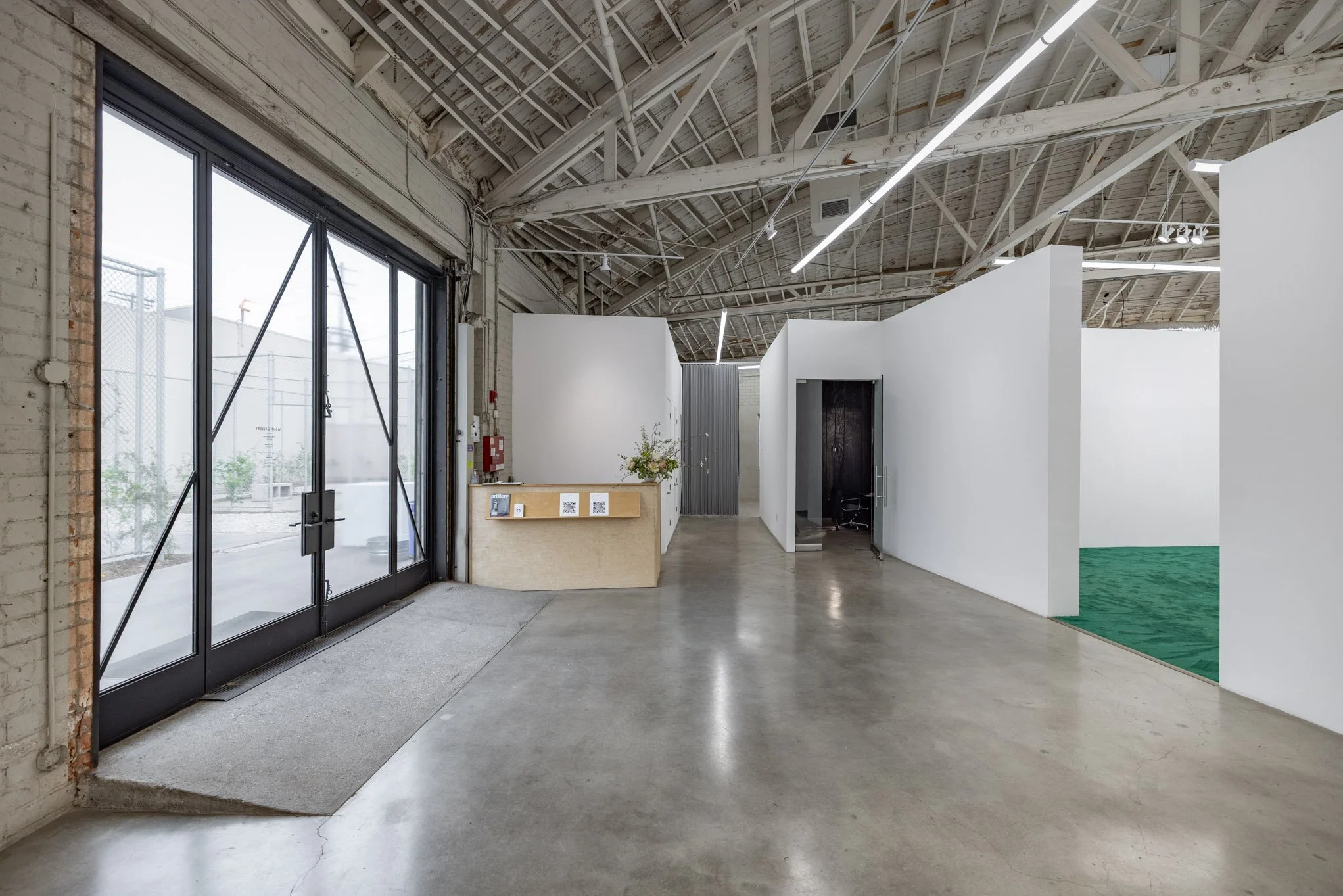 Interior view of an art gallery or showroom with white partition walls, concrete floor, and exposed ceiling with structural beams and linear lights.