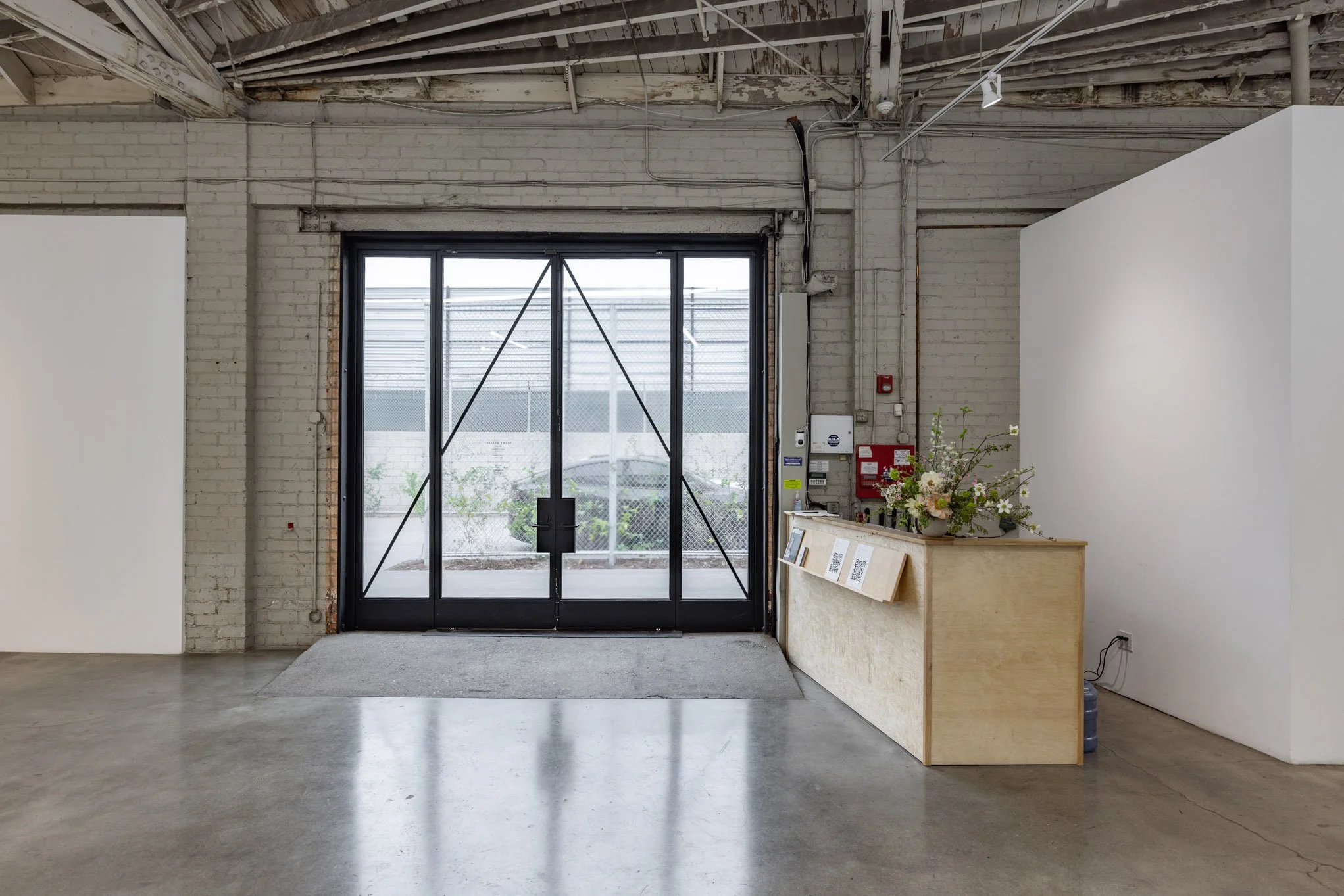 Interior space with a glass door, white brick walls, exposed ceiling pipes and lighting, and a wooden counter with a vase of flowers and papers attached.
