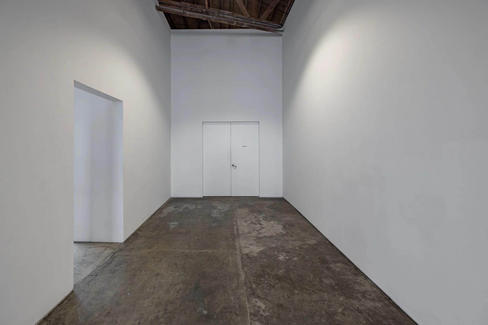 Empty white-walled room with a double door at the far end, open doorway on the left, exposed wooden ceiling, and a concrete floor.